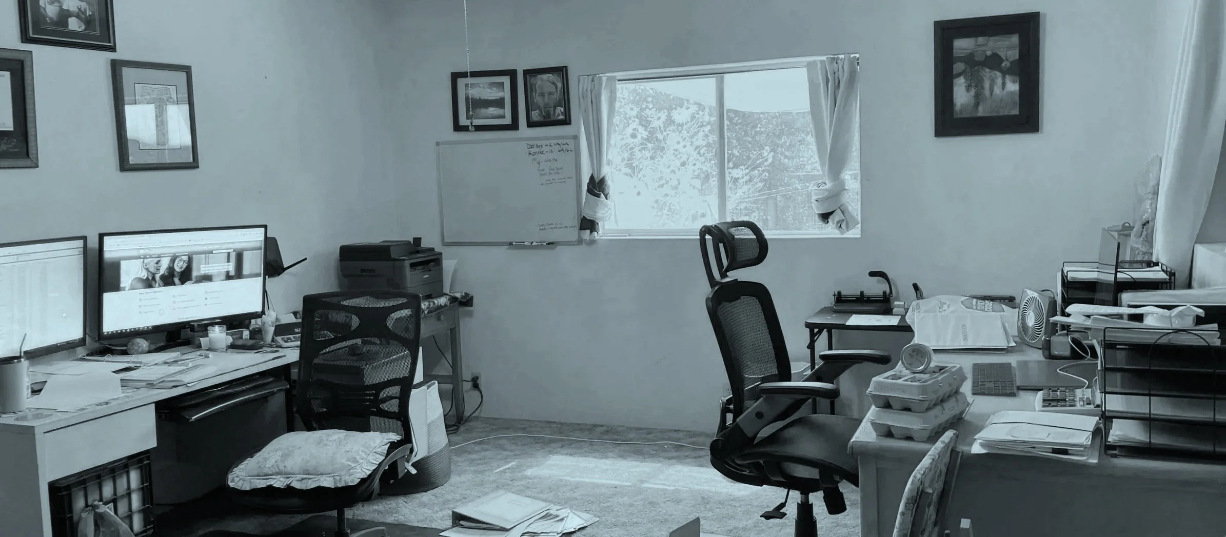 A cluttered home office with desks, office chairs, computers, stacks of paper, and office supplies. There are framed pictures on the walls and a window with curtains letting in natural light.