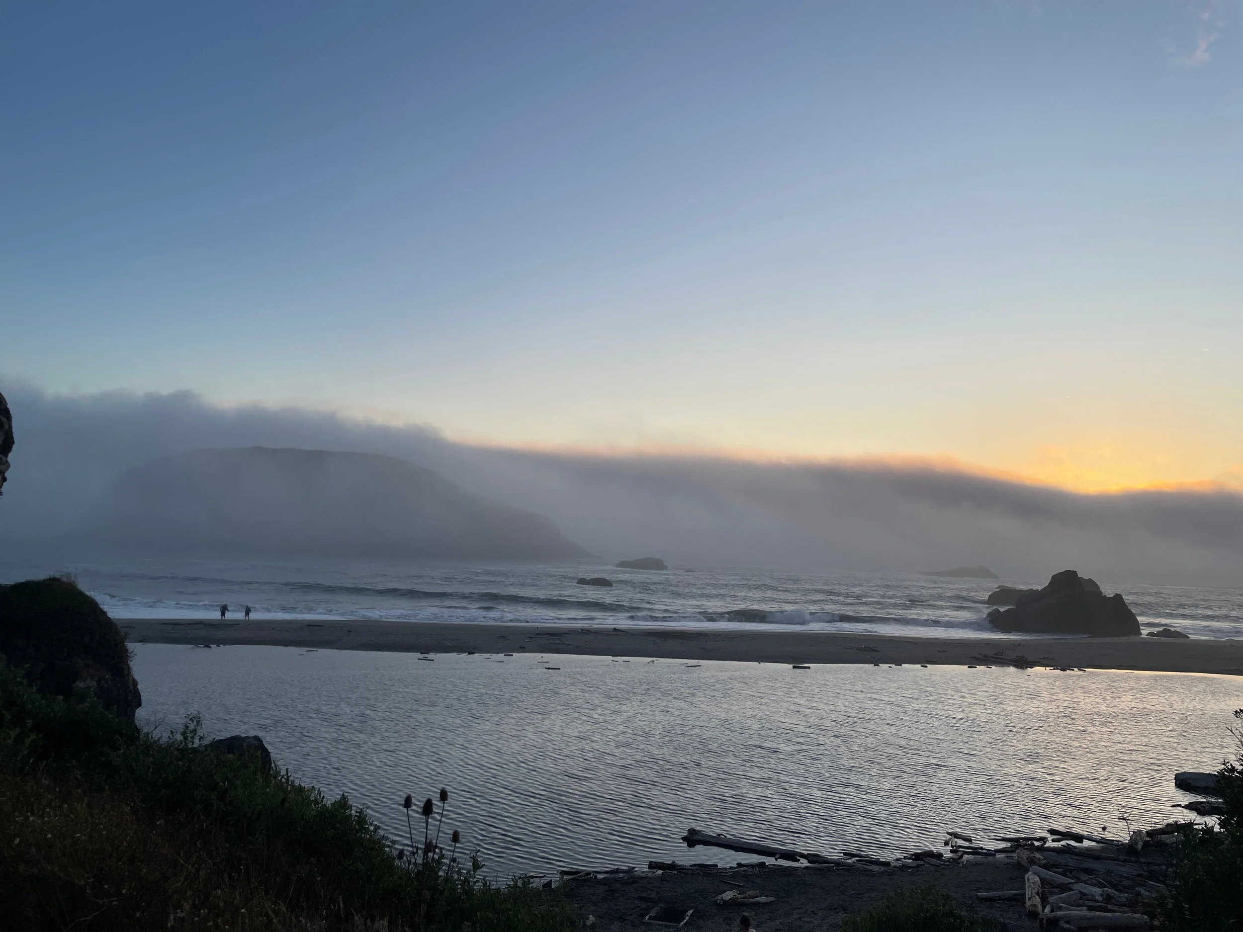 A coastal scene at sunset with a cloudy sky, ocean waves, rocky formations, and two people walking on the beach.