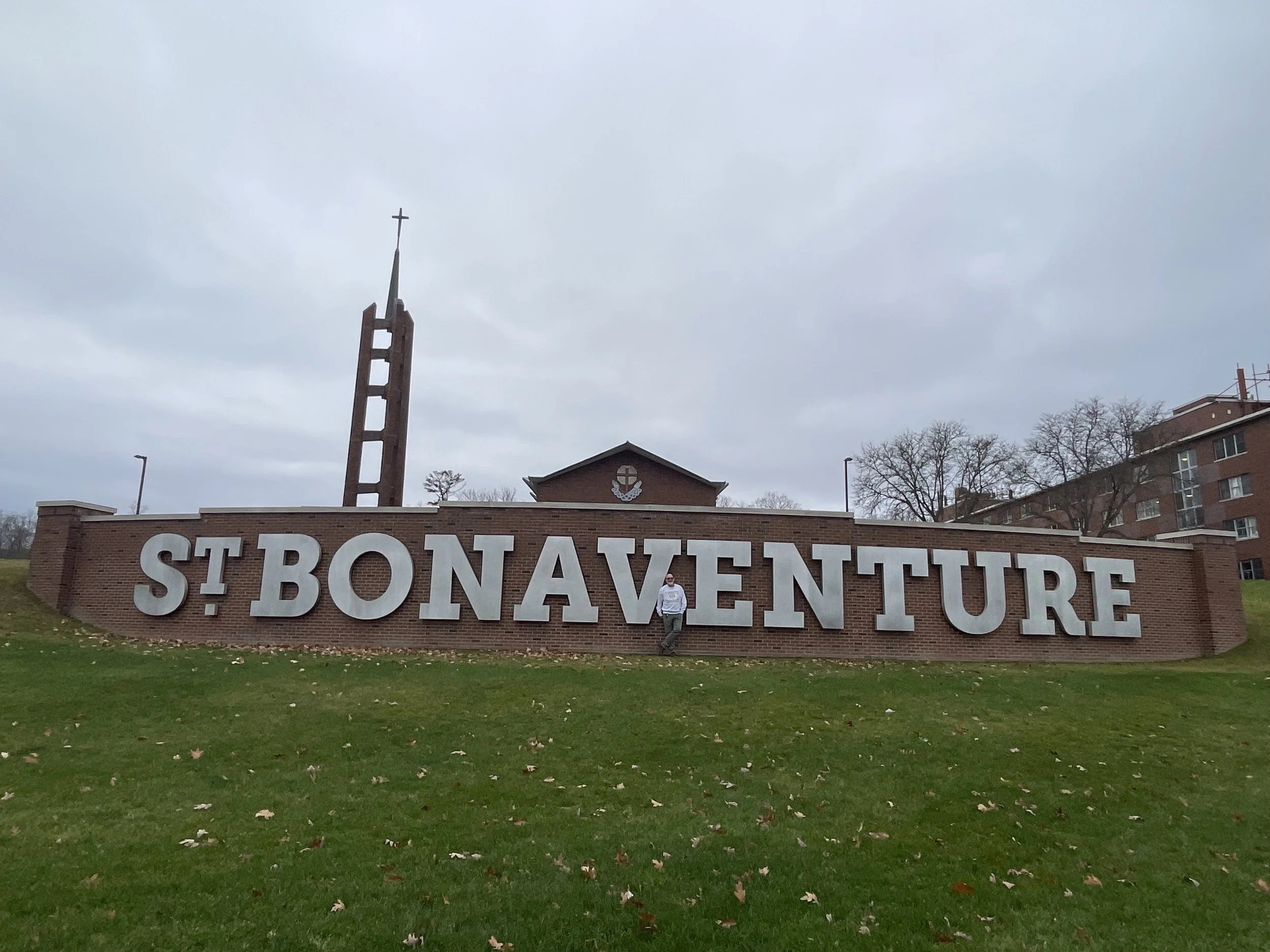 Entrance sign for St. Bonaventure University with large white letters on a brick wall, a church steeple with a cross, and buildings in the background under cloudy skies.