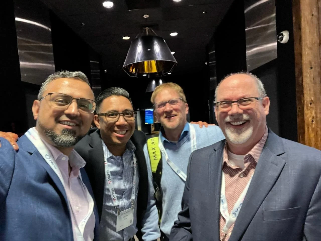 Four smiling men in professional attire posing for a photo at a corporate event or conference, with black and wooden interior background and hanging ceiling lights.