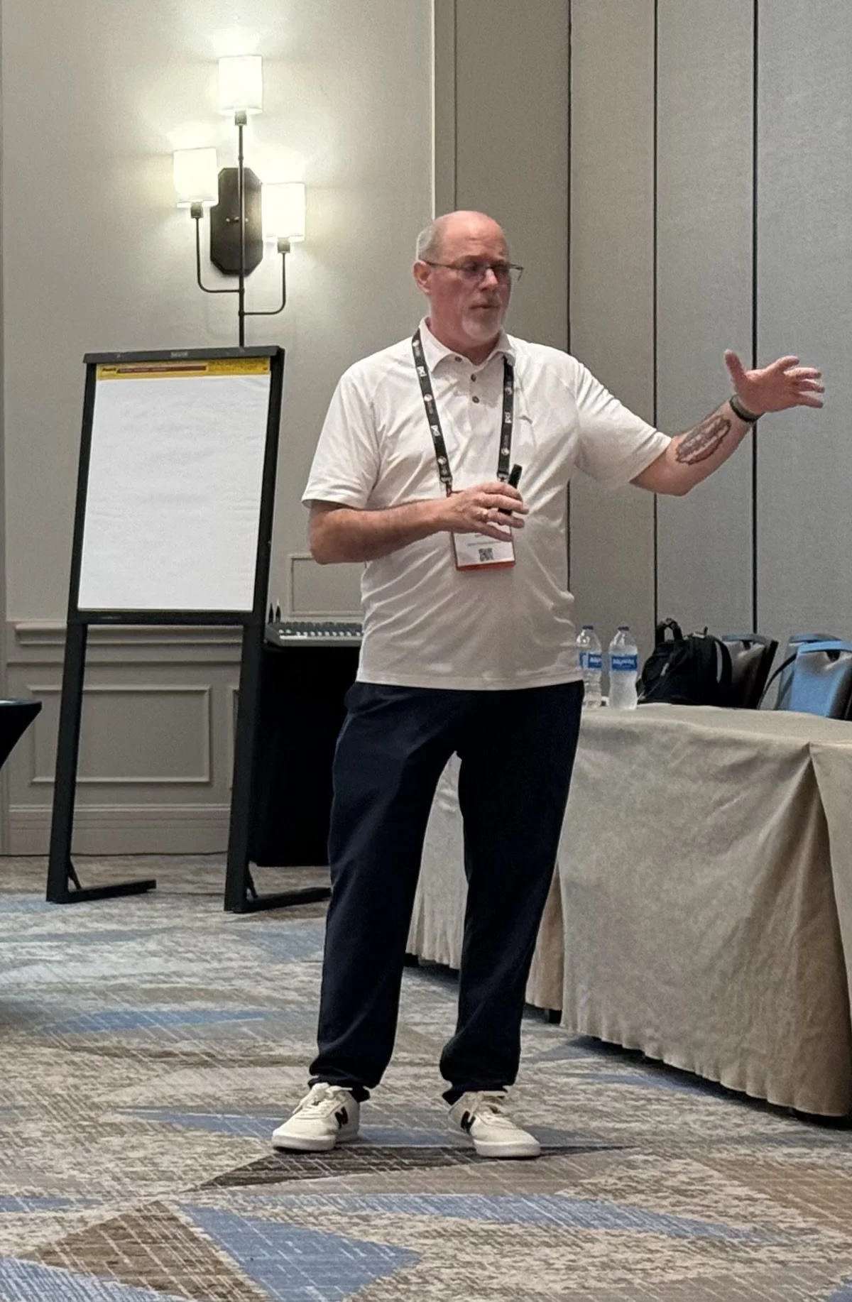 A man giving a presentation in a conference room, holding a microphone, wearing a white polo shirt, dark pants, and white sneakers, with water bottles on the table nearby.