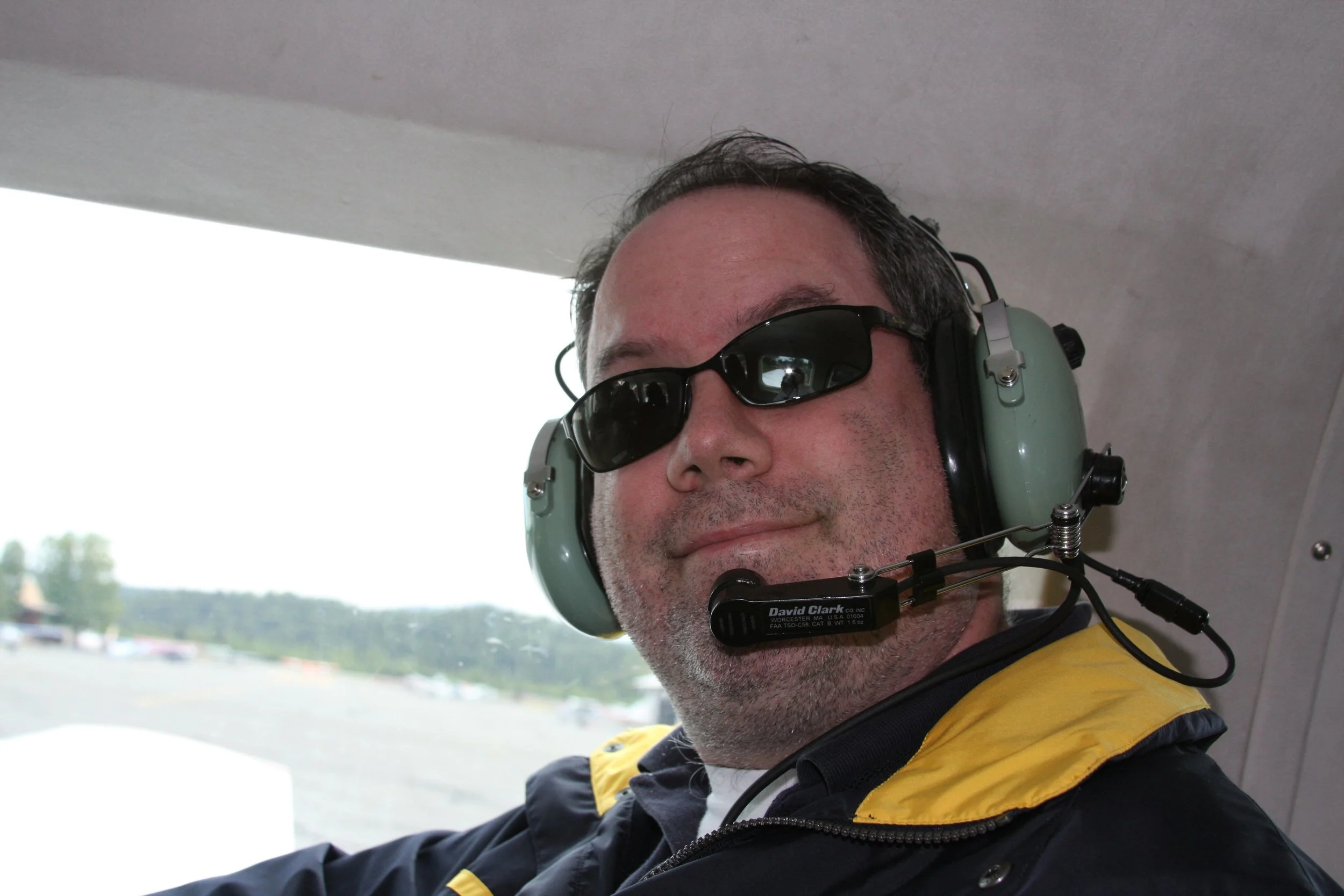 A man wearing sunglasses and a headset with a microphone, smiling inside an aircraft.