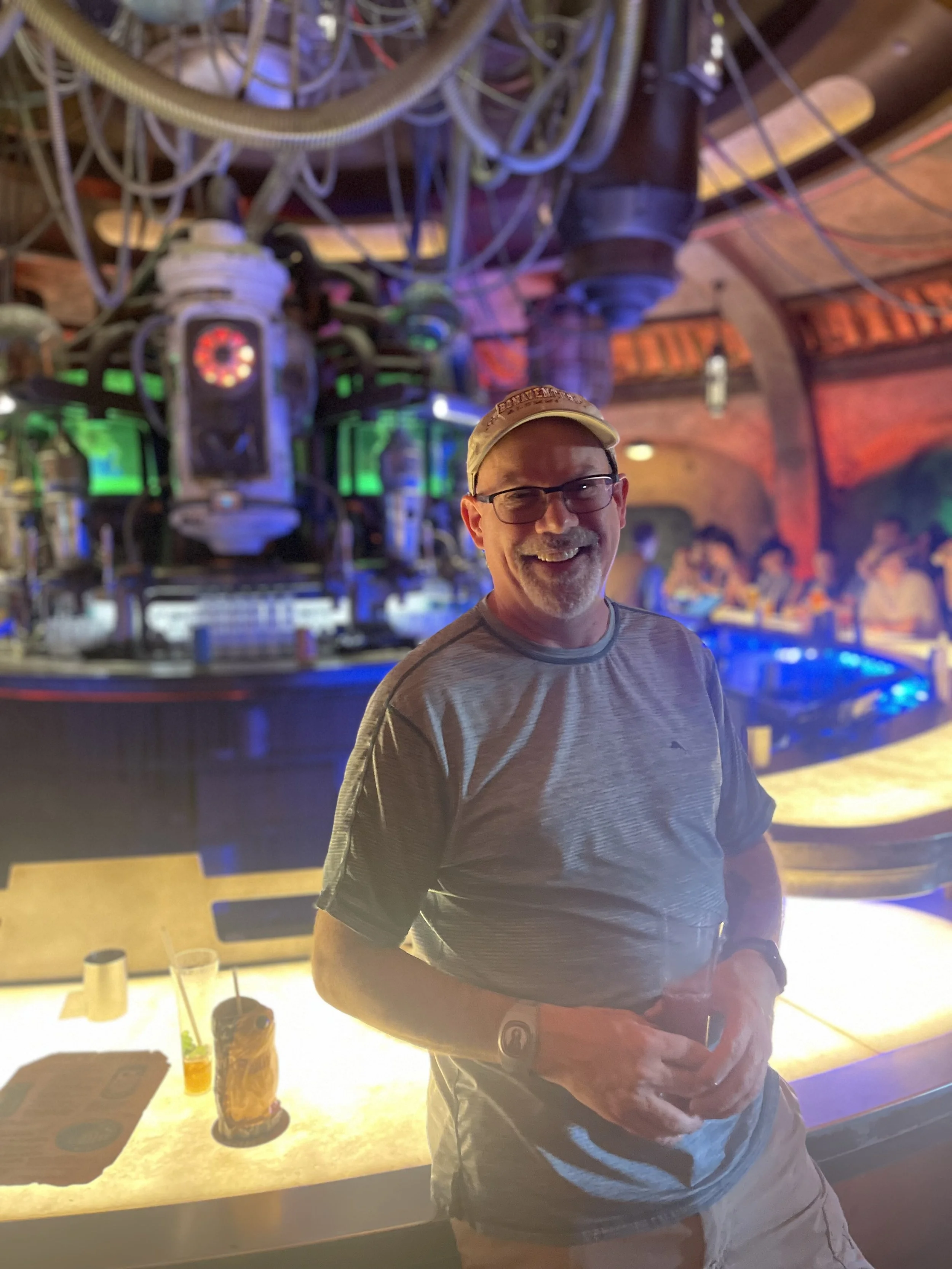 A smiling man wearing glasses and a baseball cap, standing at a bar with colorful, sci-fi themed background and patrons in a lively, futuristic bar or club.