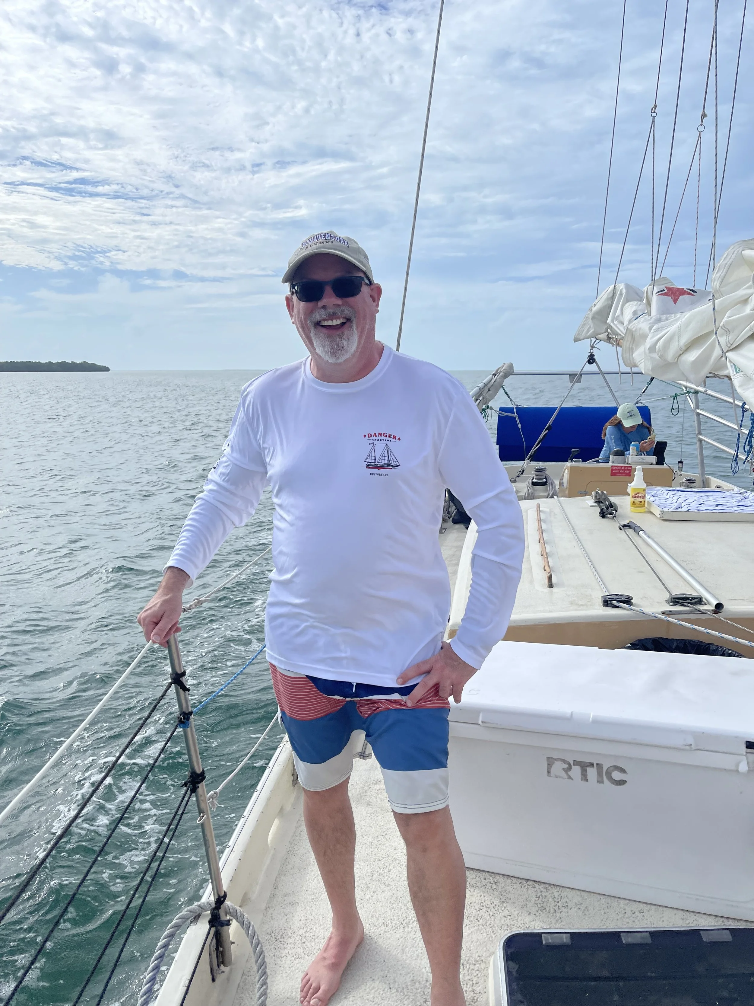 A man smiling on a sailboat, wearing sunglasses, a light-colored baseball cap, a white long-sleeve shirt, and colorful shorts, standing barefoot near the side of the boat with the ocean and cloudy sky in the background.