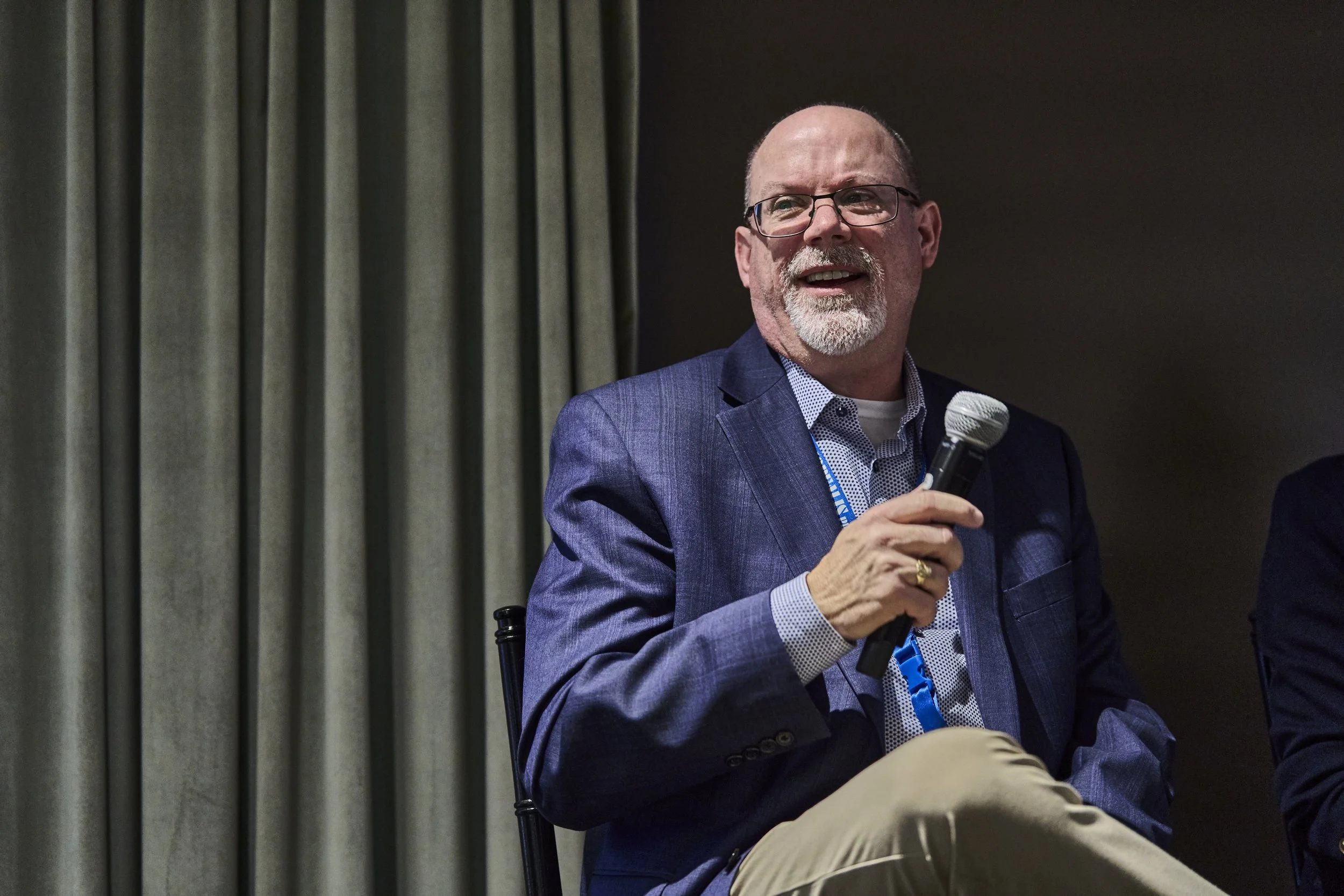 A man with glasses and a white beard holding a microphone, dressed in a blue blazer and light-colored pants, sitting on a chair against a background of curtains.