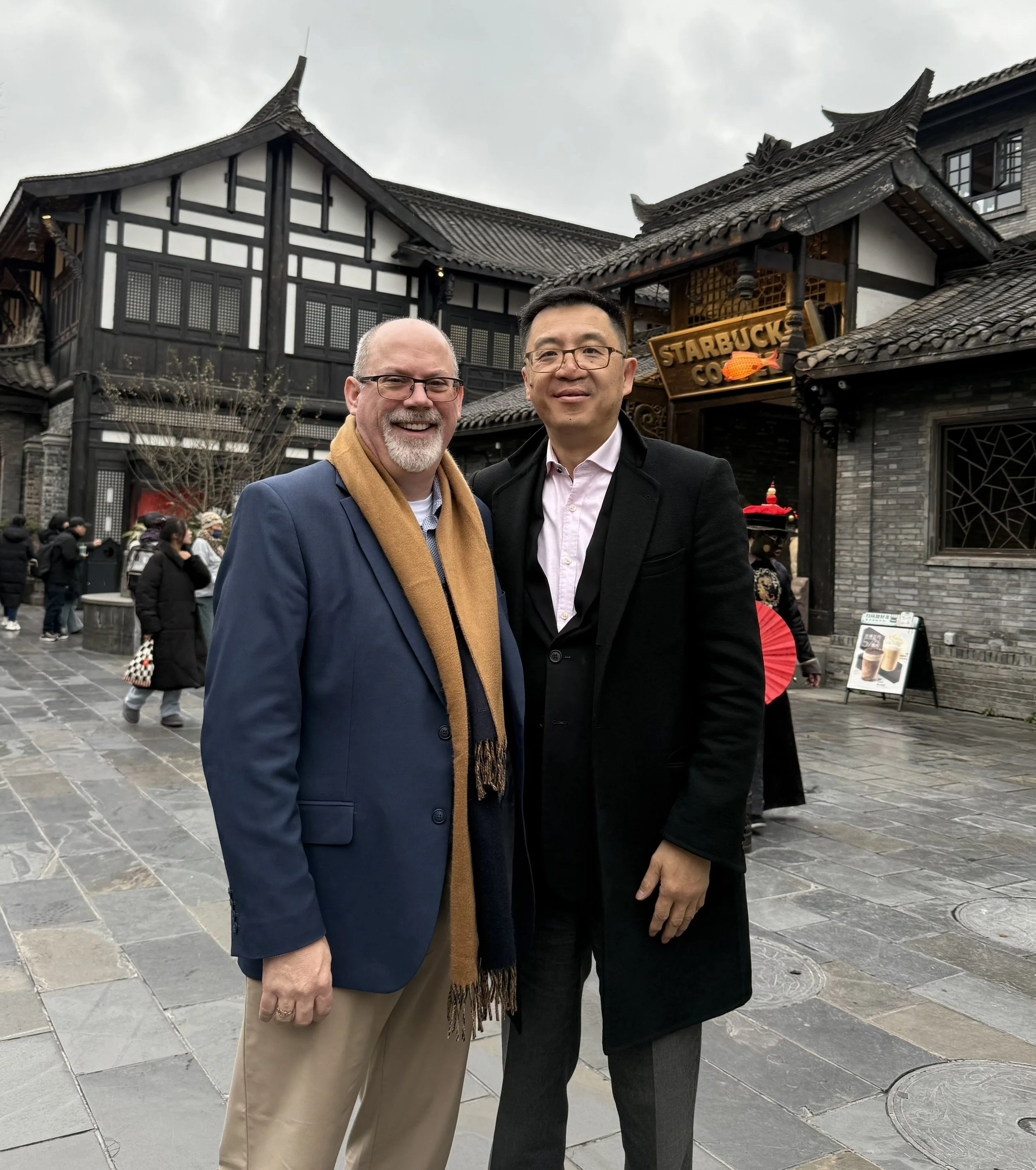 Two men standing together in front of a traditional Asian-style building with a Starbucks sign. The man on the left is smiling, wearing glasses, a blue blazer, and a tan scarf. The man on the right, also smiling, is wearing glasses, a black coat, and a light pink shirt. There are several people in the background walking on a paved area under an overcast sky.