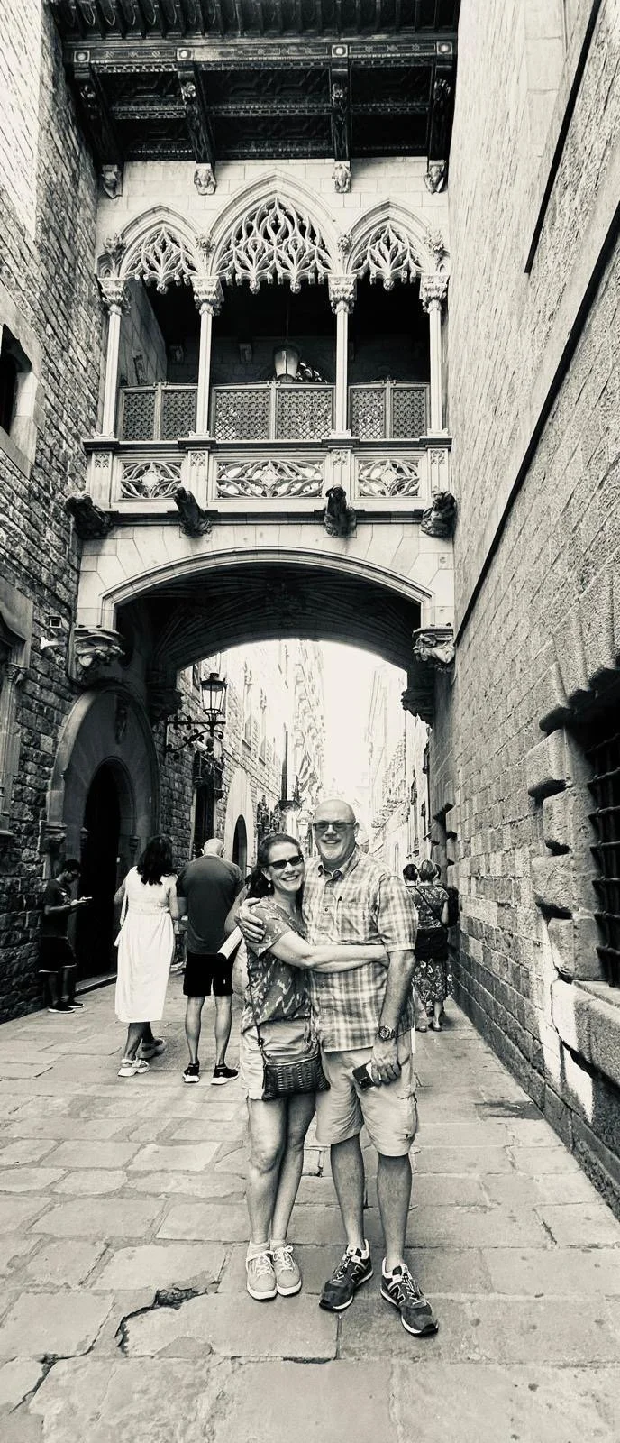 A black and white photo of a couple smiling and hugging on a narrow historic street with stone buildings, arched bridge, and decorative architectural features.