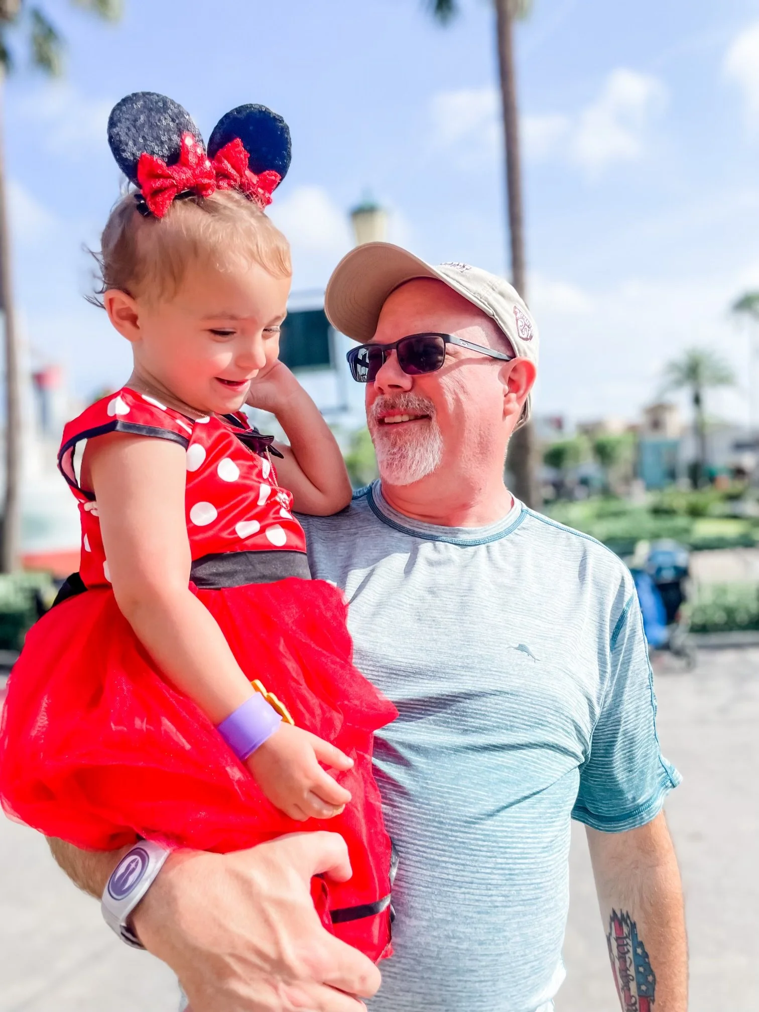 A young girl dressed as Minnie Mouse being held by a man at an outdoor amusement park or Disney theme park on a sunny day.