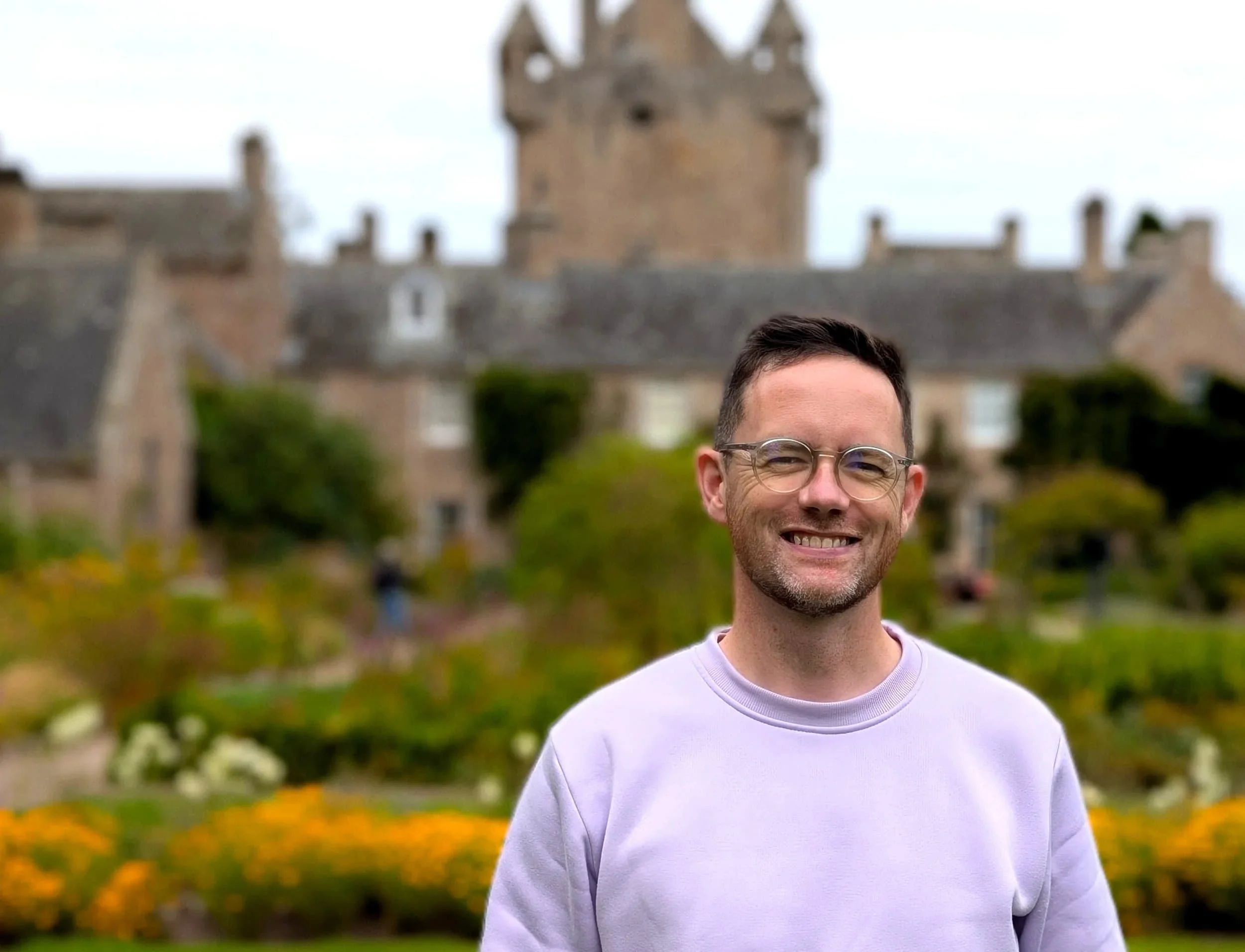 A smiling man with glasses and short dark hair stands outdoors in front of a historic stone castle with a garden. The sky is cloudy.