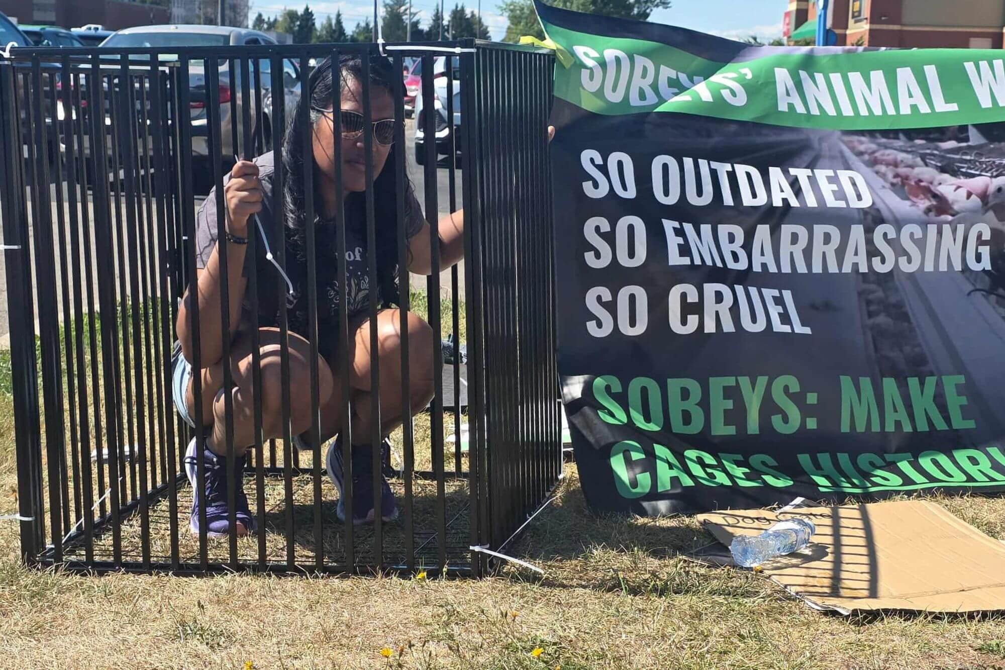 A woman squatting inside a cage at a protest, holding a small object in her right hand. To her right is a large black and green banner with white and green text protesting animal cruelty and urging to make cages history.