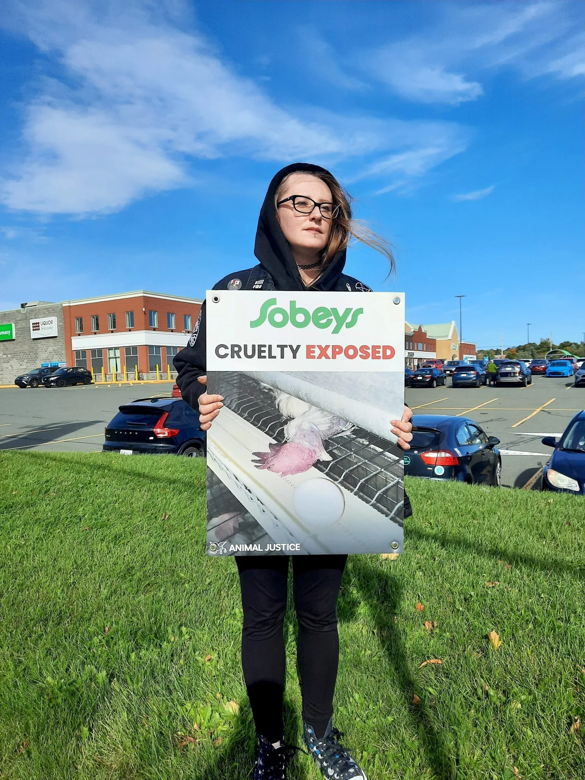 A woman with long hair, glasses, and a choker holding a sign that says 'Sobeys Cruelty Exposed' with an image of a baby chicken behind a wire.