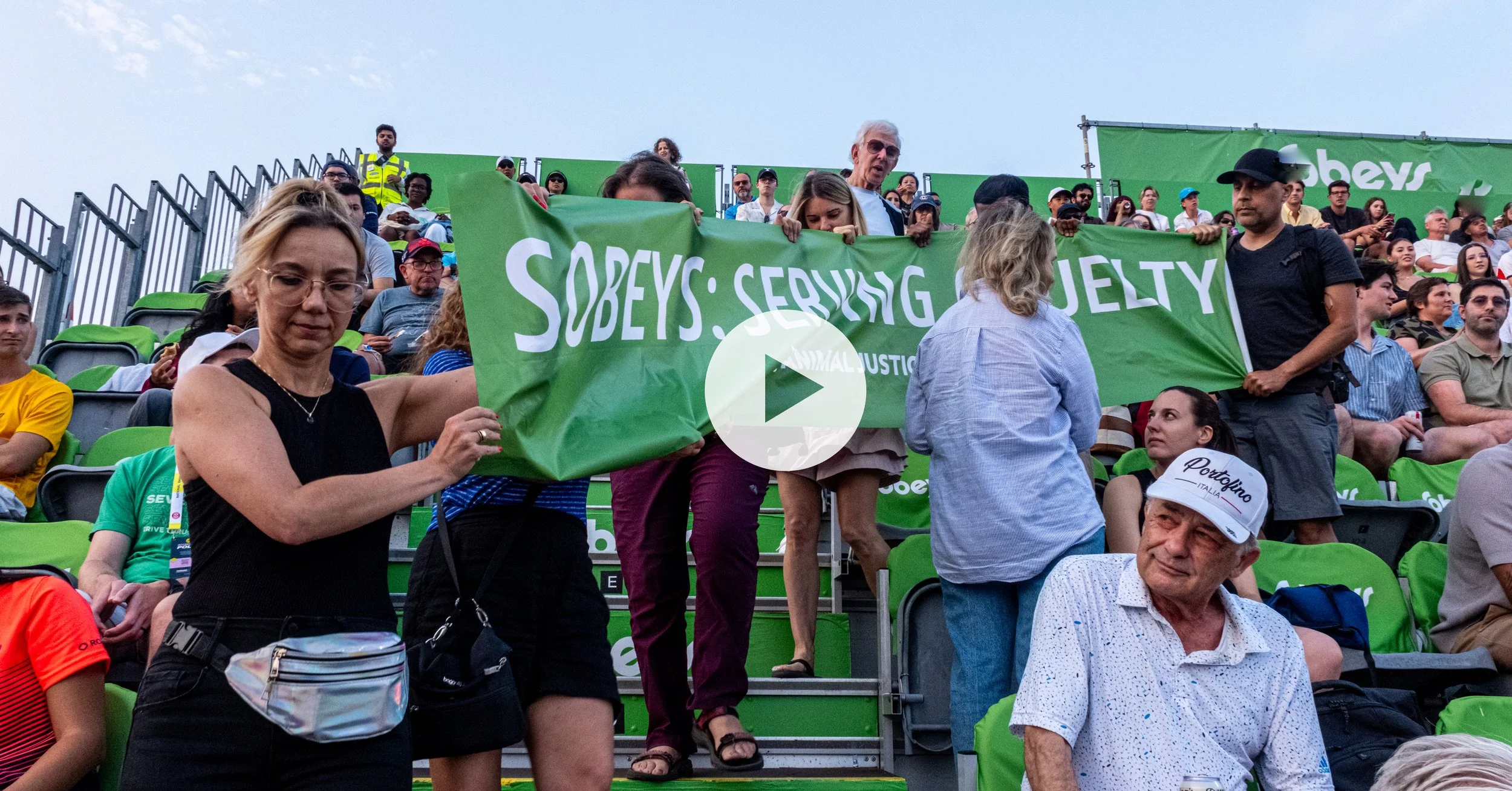 People holding a green banner at a protest or rally with a crowd seated in stadium seats in the background.