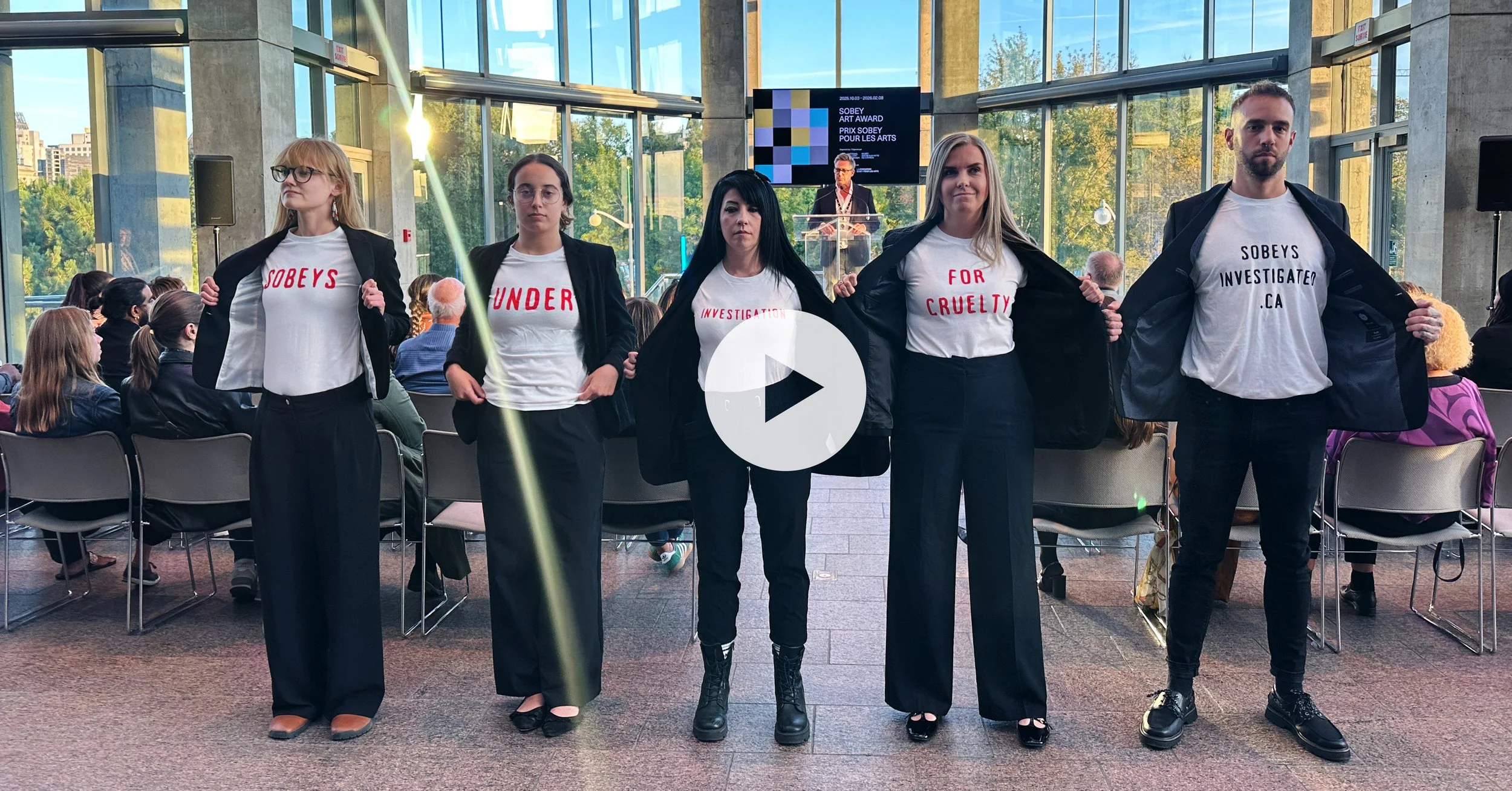 Group of five protesters with opened black jackets revealing protest slogans on white shirts inside a glass-walled building, with an audience watching in the background.