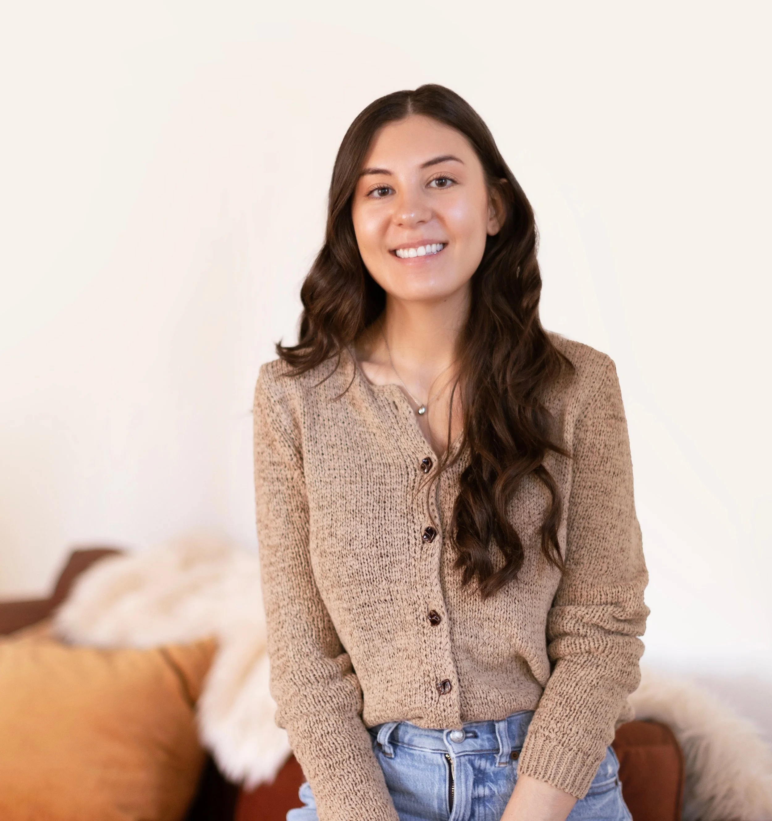 A young woman with long, wavy brown hair and a tan cardigan smiling in a room with a white wall and a brown couch with pillows in the background.