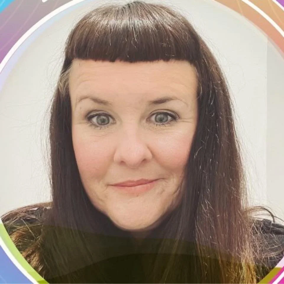 Close-up of a woman with dark brown hair styled with blunt bangs, blue eyes, and a slight smile, against a colorful background.