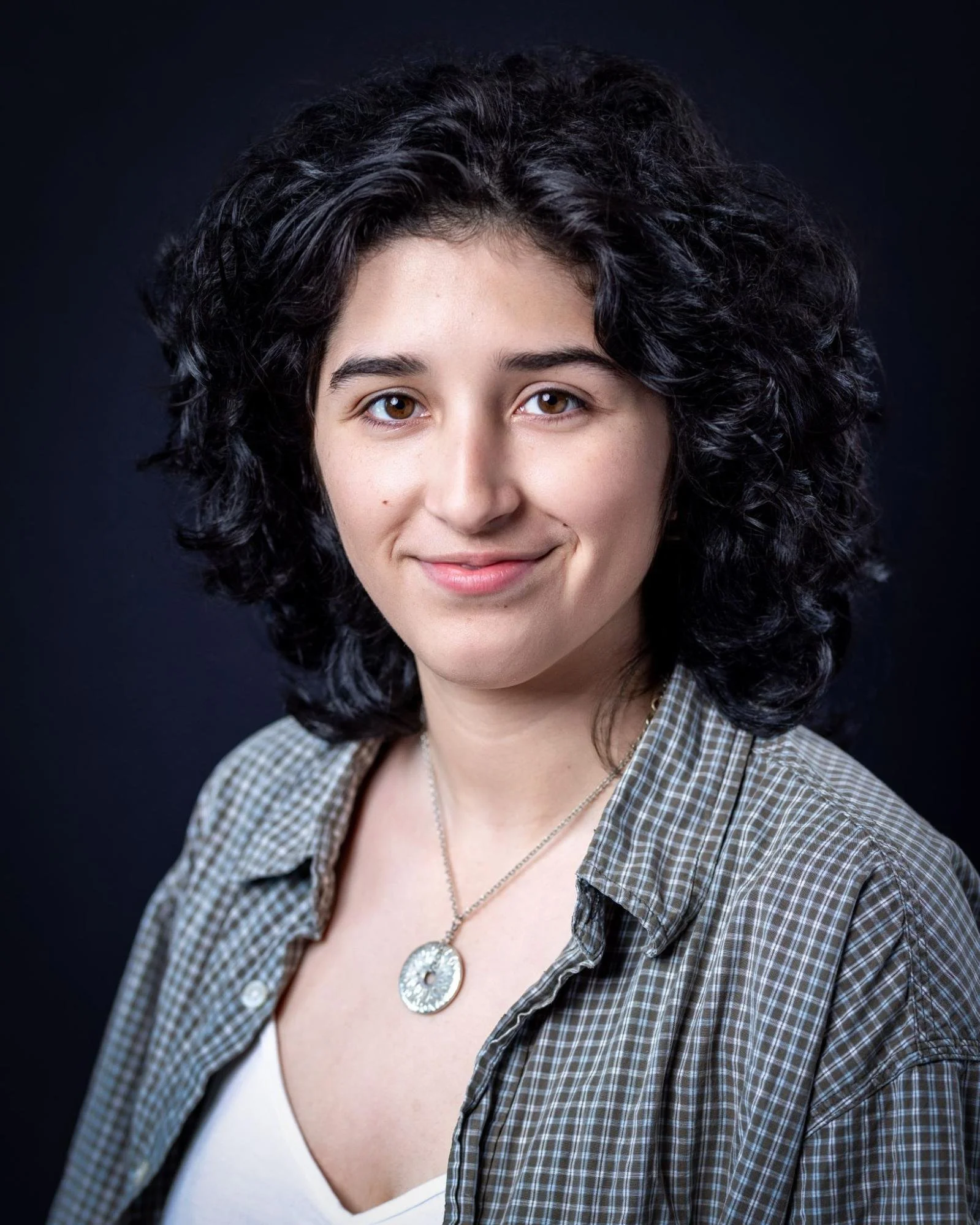 Portrait of a woman with curly dark hair, wearing a checkered shirt and a necklace, smiling against a dark background.