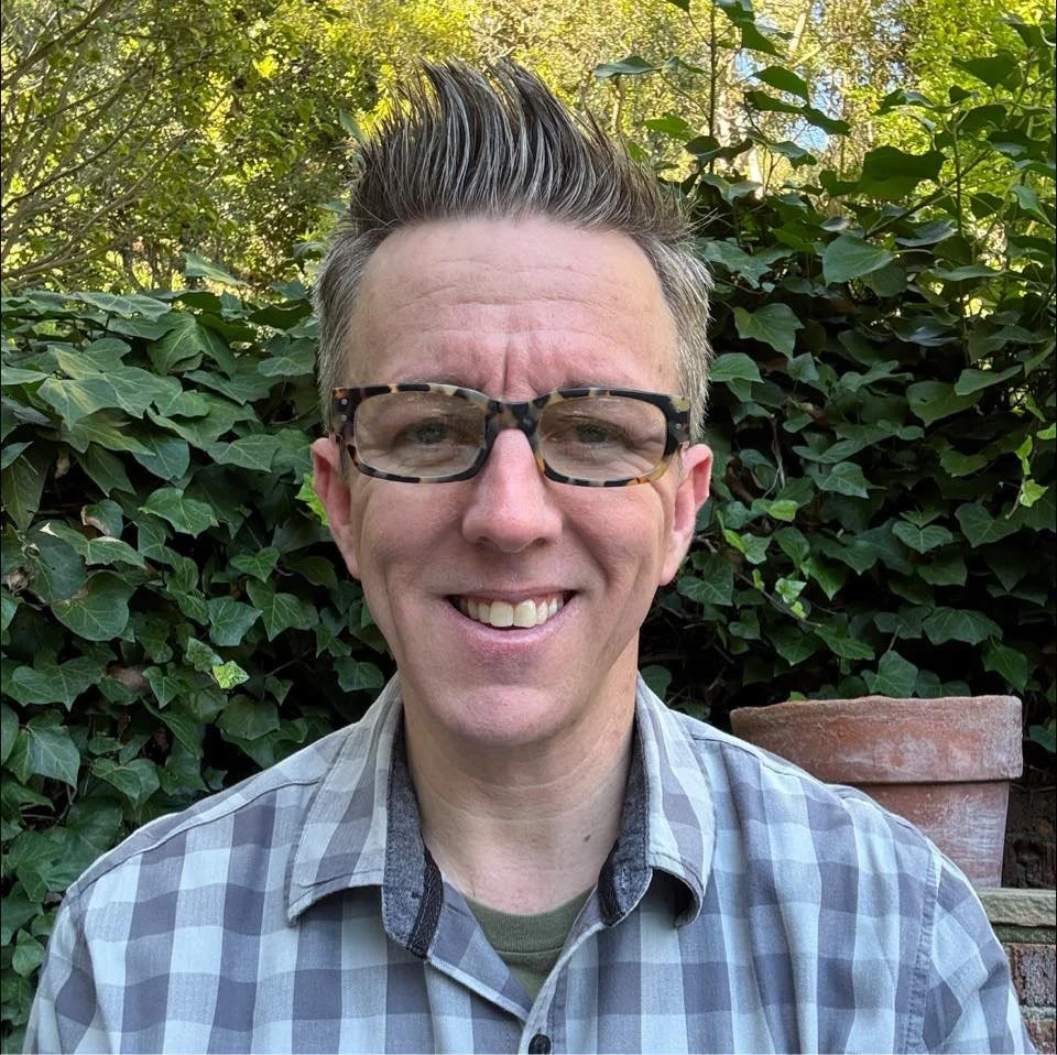 A smiling man with spiked hair and glasses, wearing a plaid shirt, standing outdoors in front of leafy green plants and a brick wall with a flowerpot.