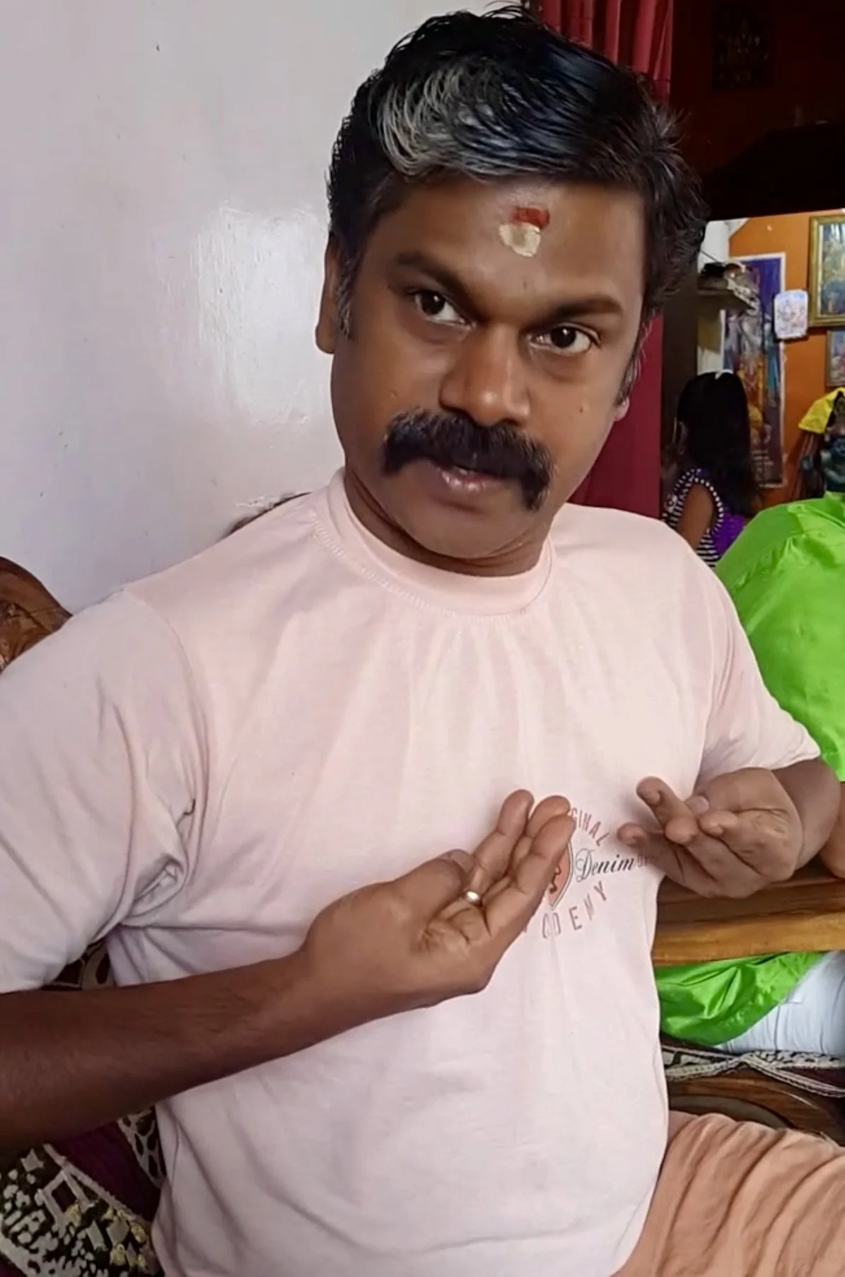 A man with dark hair, a mustache, and a tilak on his forehead, making a gesture with his hand in a casual indoor setting.