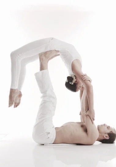 Two women practicing acro yoga, one lying on her back and lifting the other above her using her legs, both dressed in white against a plain white background.
