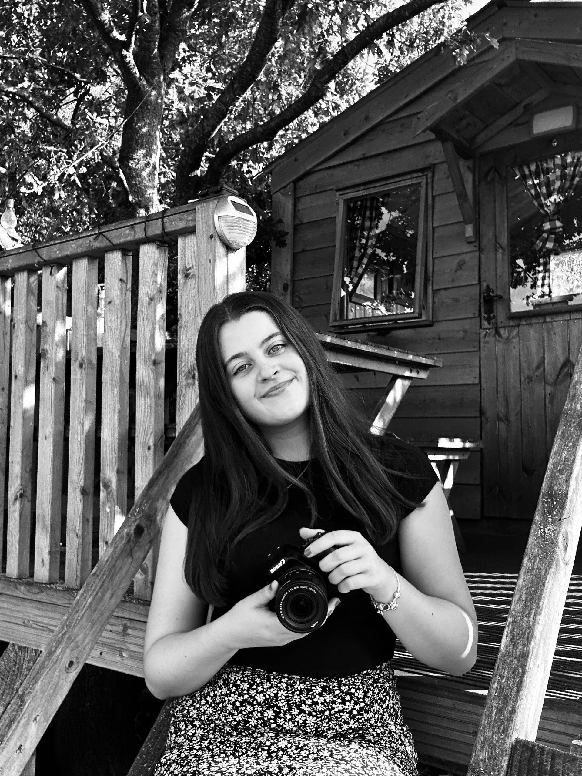 A smiling young woman with long dark hair holding a camera, standing on a wooden deck outdoors with a small wooden shed and trees in the background.