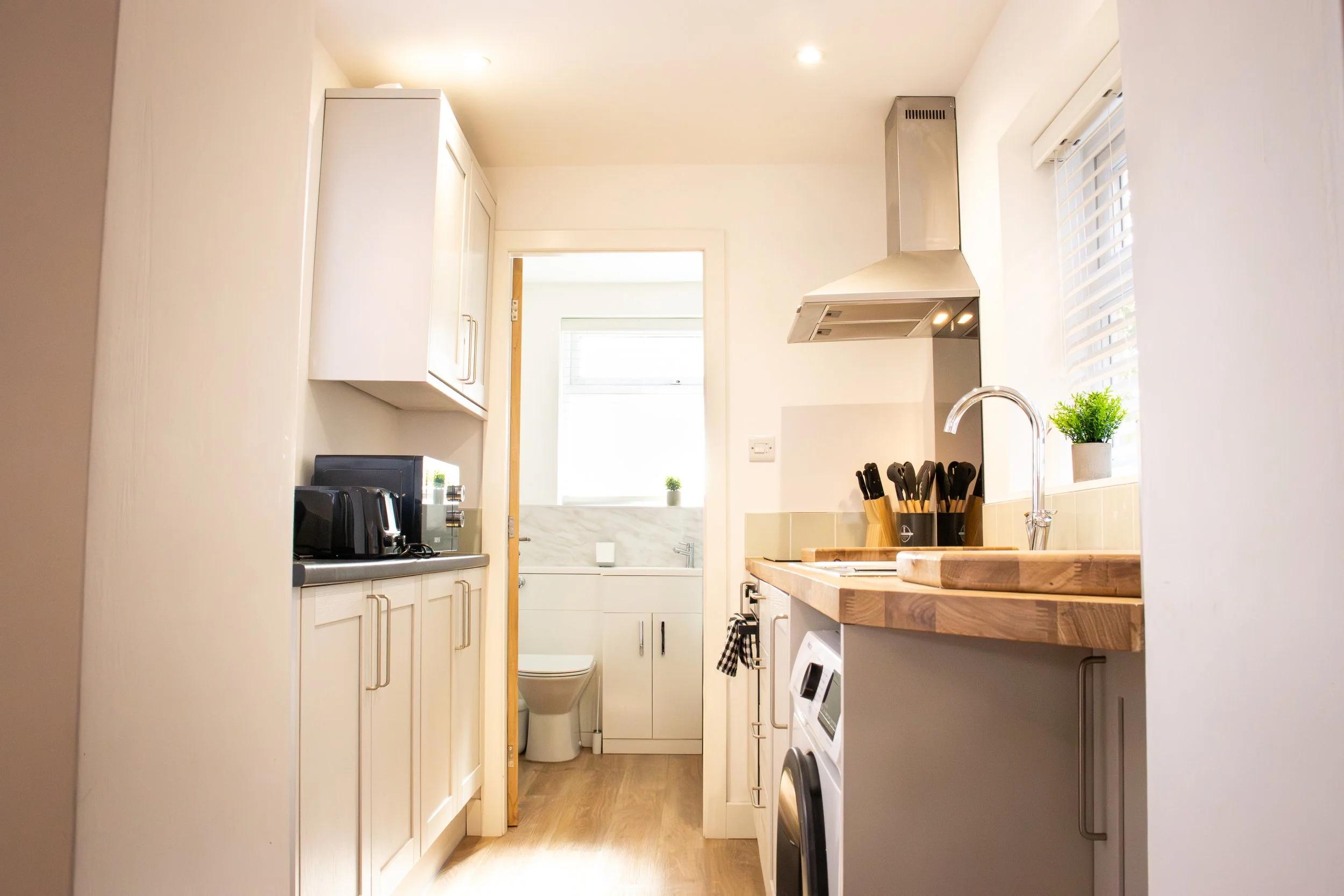 A compact kitchen with white cabinets, a wooden countertop, a stainless steel range hood, a small window with blinds and a potted plant, and a view into a small bathroom with a toilet and a window.