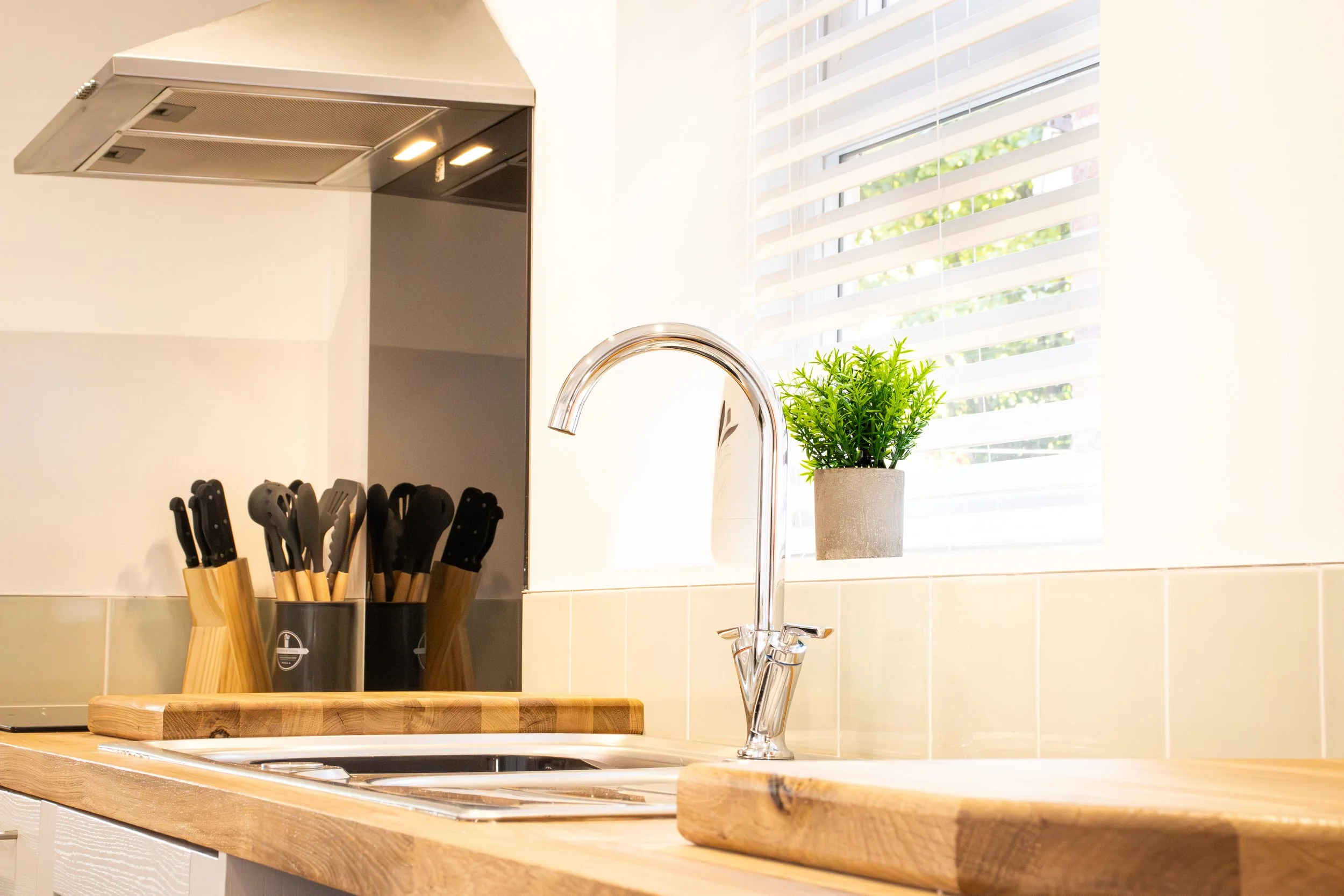 A kitchen sink with a chrome faucet, a wooden cutting board, a potted green plant on a windowsill, and cooking utensils in black holders on a light-colored wooden countertop.