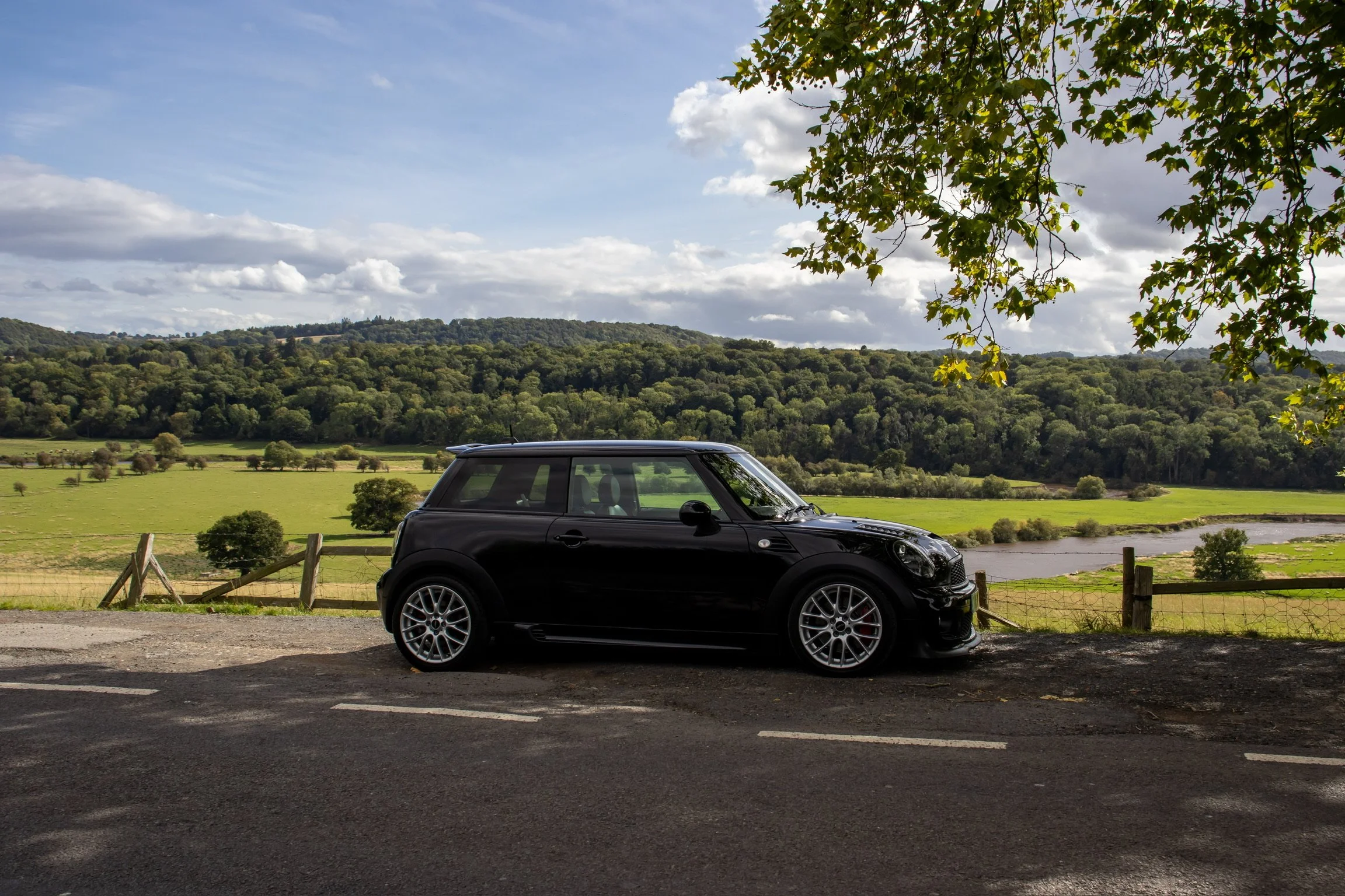 Black Mini Cooper parked on side of rural road with a green field, trees, a river, rolling hills, and cloudy sky in background.