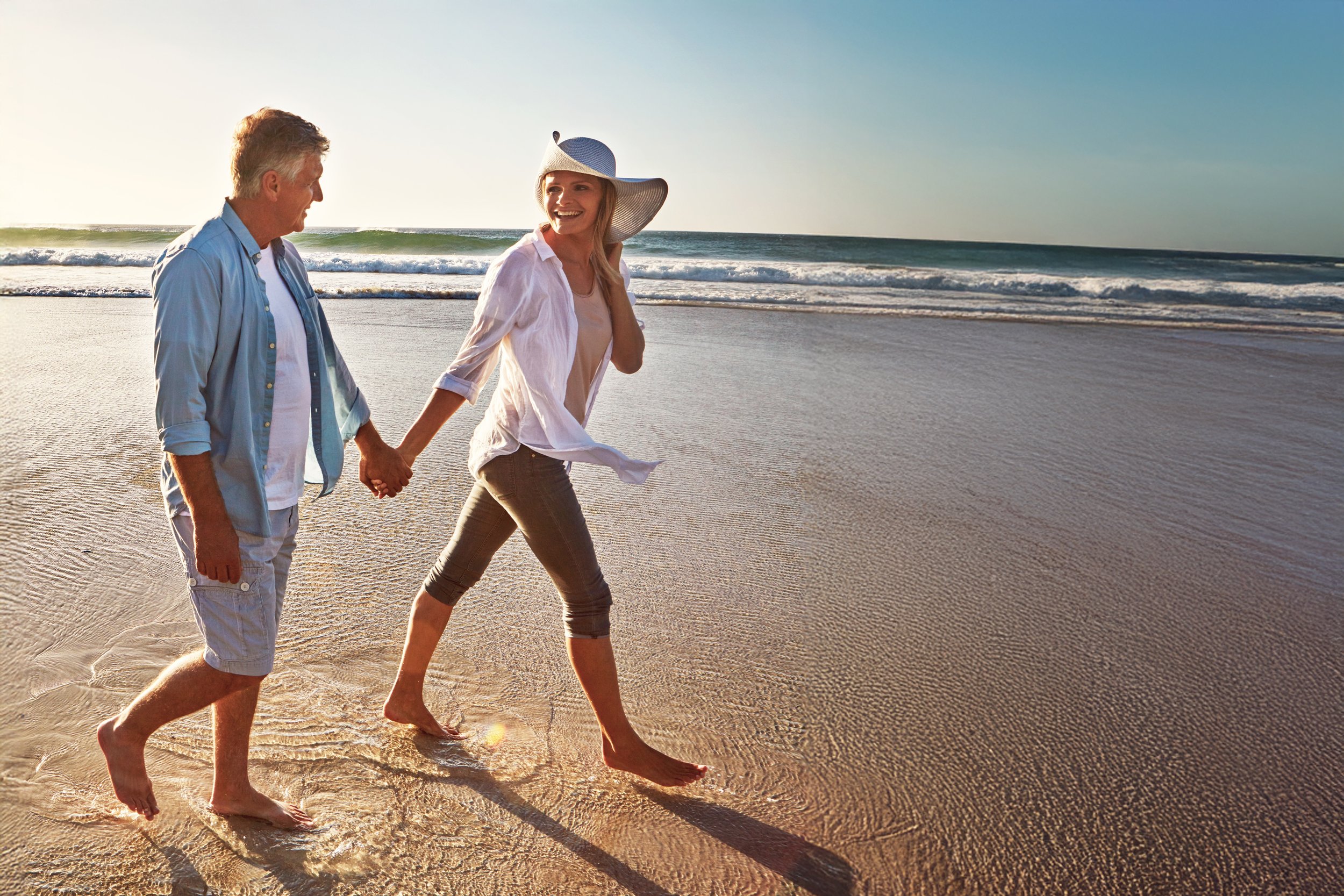 A couple holding hands and walking barefoot on the beach near the ocean during sunset, smiling and enjoying their time together.