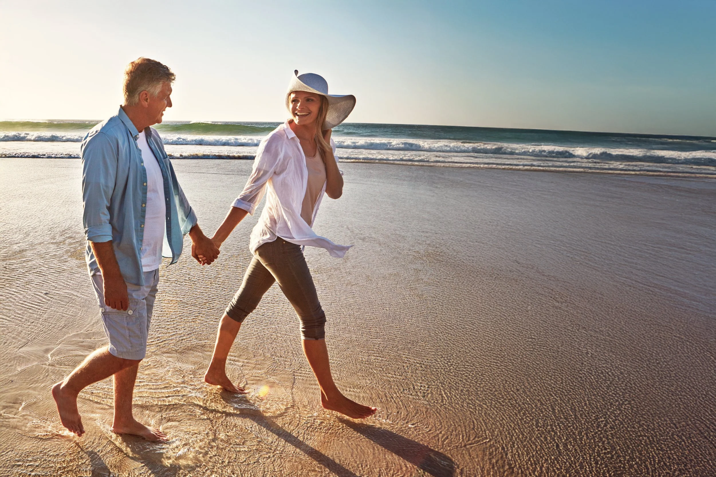 A couple holding hands and walking barefoot on the beach near the ocean during sunset, smiling and enjoying their time together.