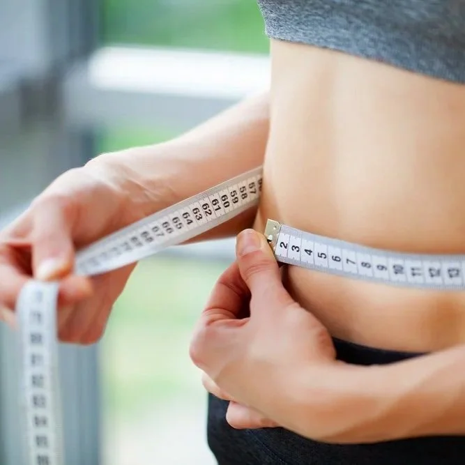 Person measuring their waist with a white measuring tape in a bright room.