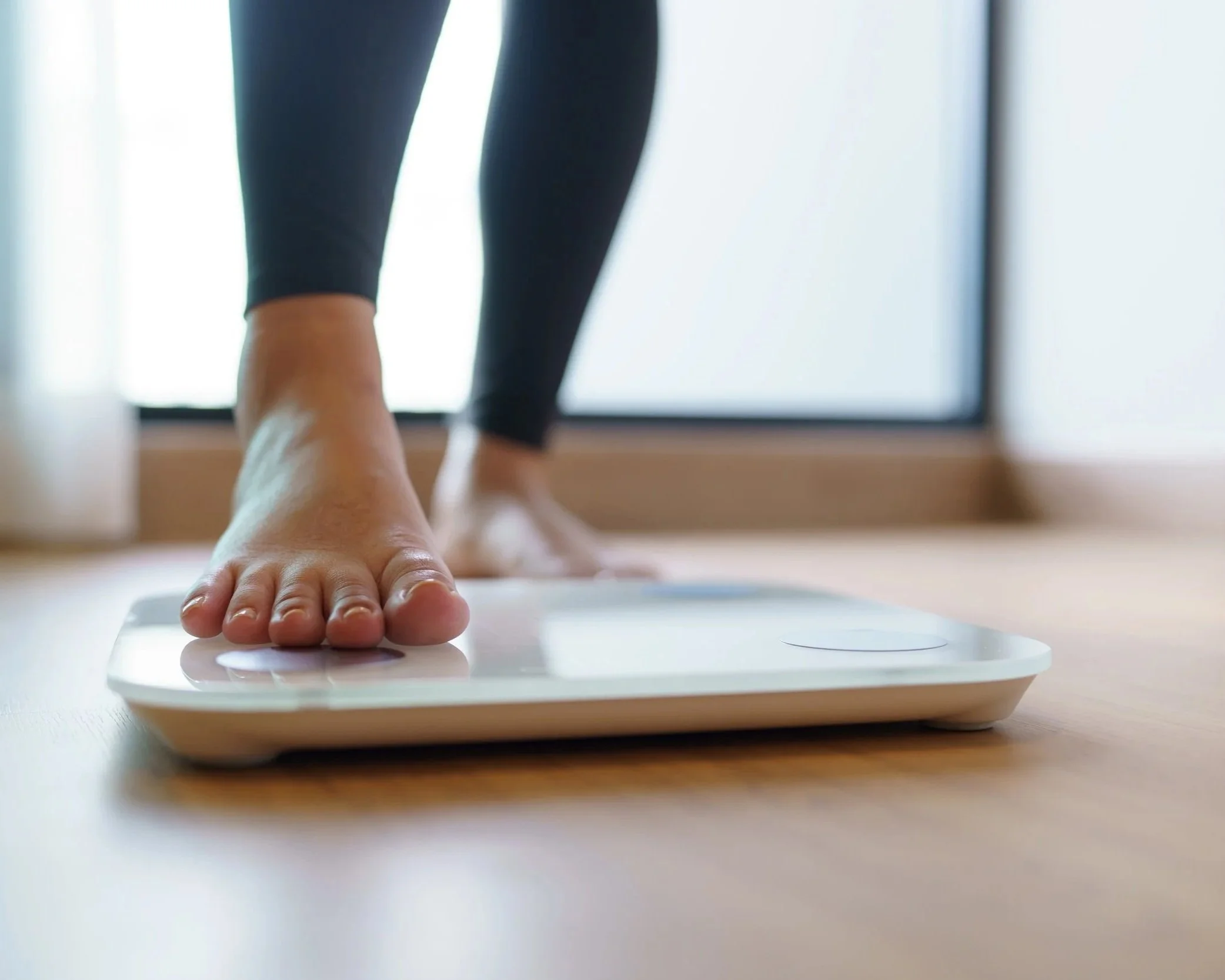A person doing yoga on a wooden floor, focusing on their hand and lower leg. The person is wearing black leggings and is positioned near a glass door or window with natural light streaming in.