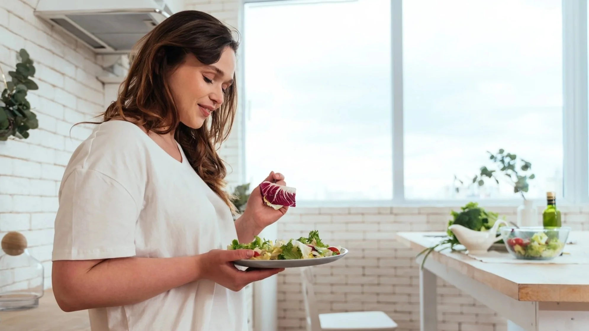 A woman with long brown hair in a white shirt holding a plate of salad, standing in a bright modern kitchen with a white brick wall and large window.