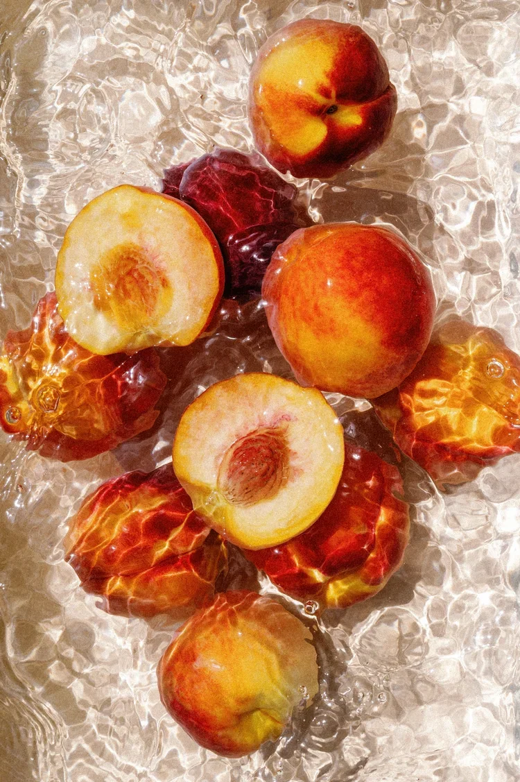 Still life editorial of ripe peaches submerged in rippling water, photographed with natural light. Captured by Natalie Folchi, known for poetic product and food photography with texture and color.