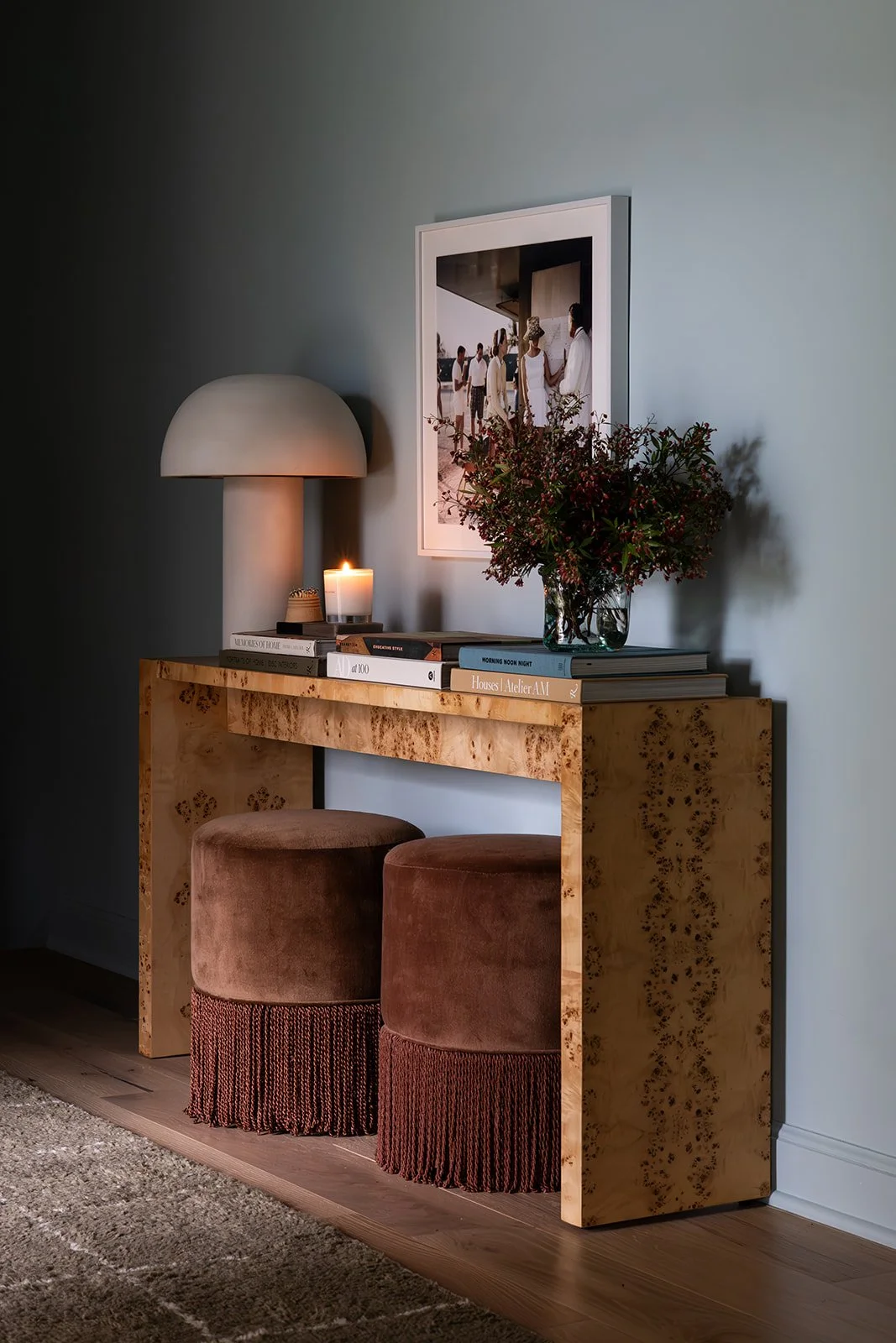 A warmly styled console table vignette featuring a burl wood console, sculptural table lamp, stacked design books, and a framed art print. Two Studio McGee velvet ottomans with fringe detailing are tucked beneath the table, while soft candlelight and