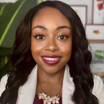 A woman with dark hair styled in loose waves, smiling and wearing a white blazer and a decorative necklace, set against a background with green plant leaves and framed pictures.