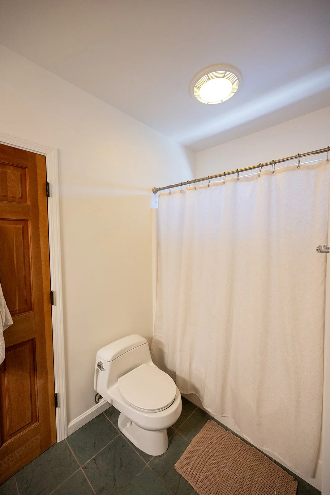A bathroom with a closed wooden door, a white toilet, a beige shower curtain on a rod, a ceiling light, a brown bath mat, and tiled floor.