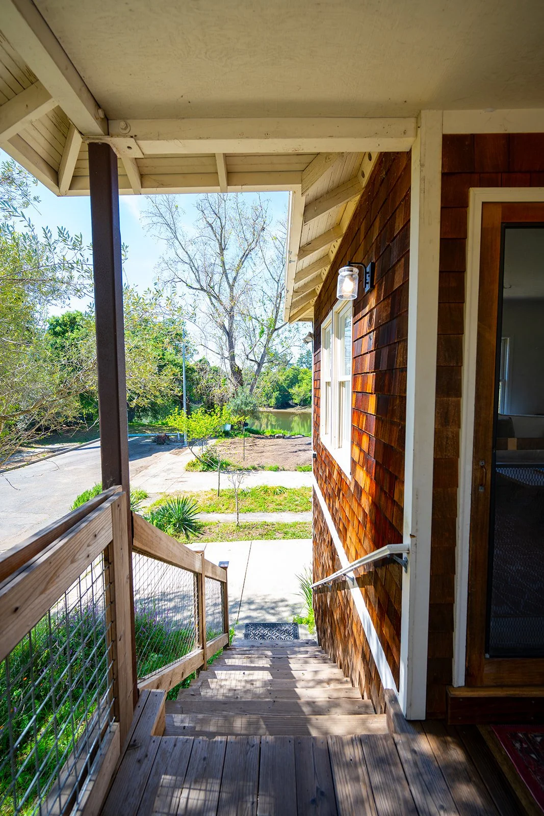 Wooden porch stairs leading down to sidewalk outside a house with brown shingle siding and a window, overlooking a garden and a tree-lined road near a body of water.