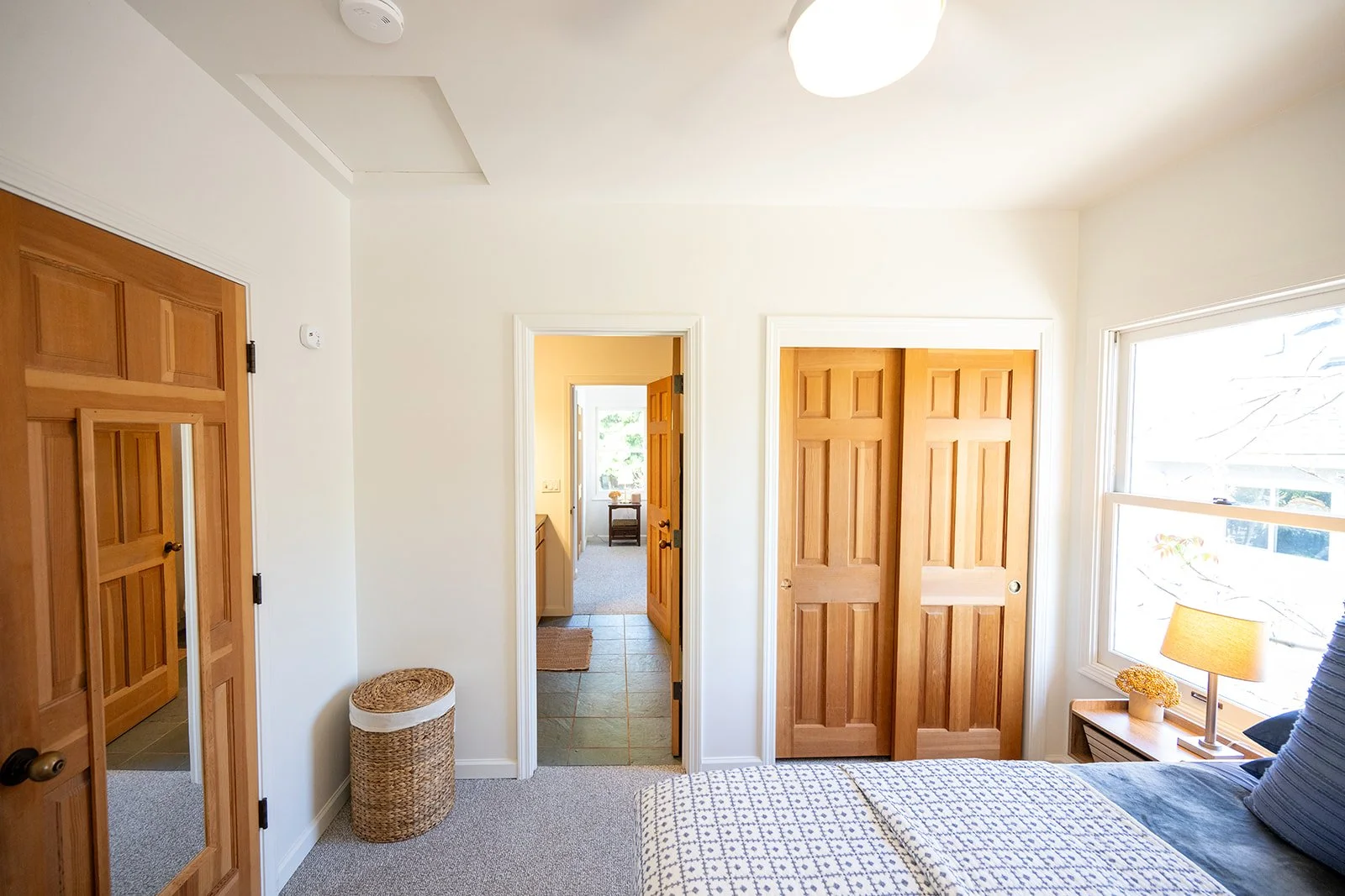 Sunlit bedroom with wooden closet doors, a bedside table with a lamp, a window, and a partially visible bed with patterned bedding.