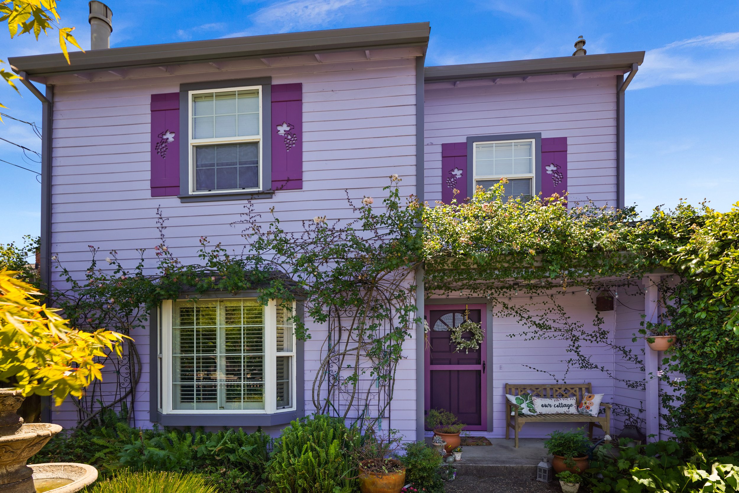 A two-story house painted in lavender with purple shutters and gray trim. There are two windows on the upper floor with purple shutters featuring decorative cutouts of grapes and leaves. The house has a small front porch with a wooden bench that has 