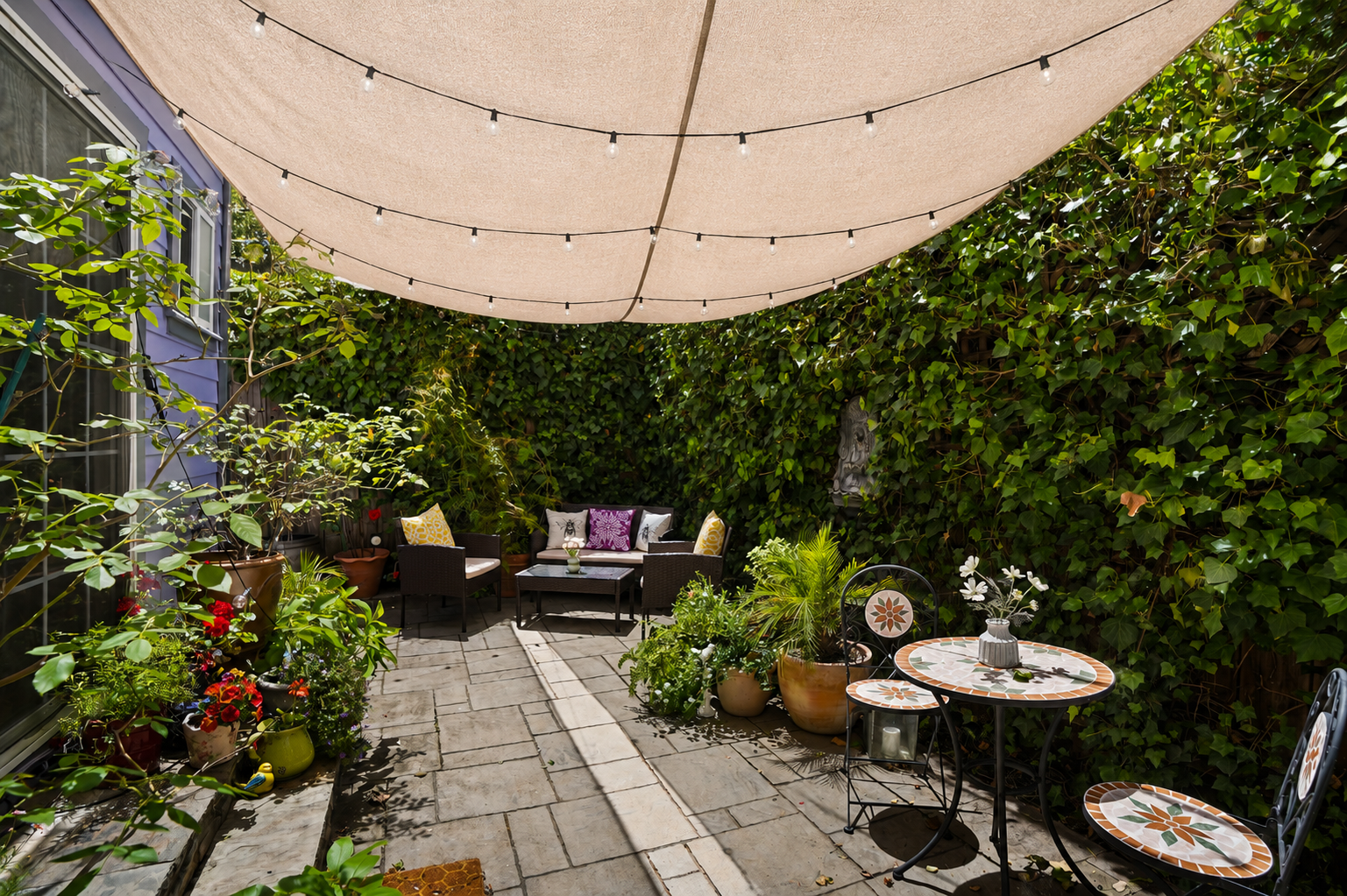 Sunny patio with a beige canopy, string lights, and potted plants. There are outdoor seating areas with colorful pillows and a tiled table with chairs, surrounded by lush green ivy.