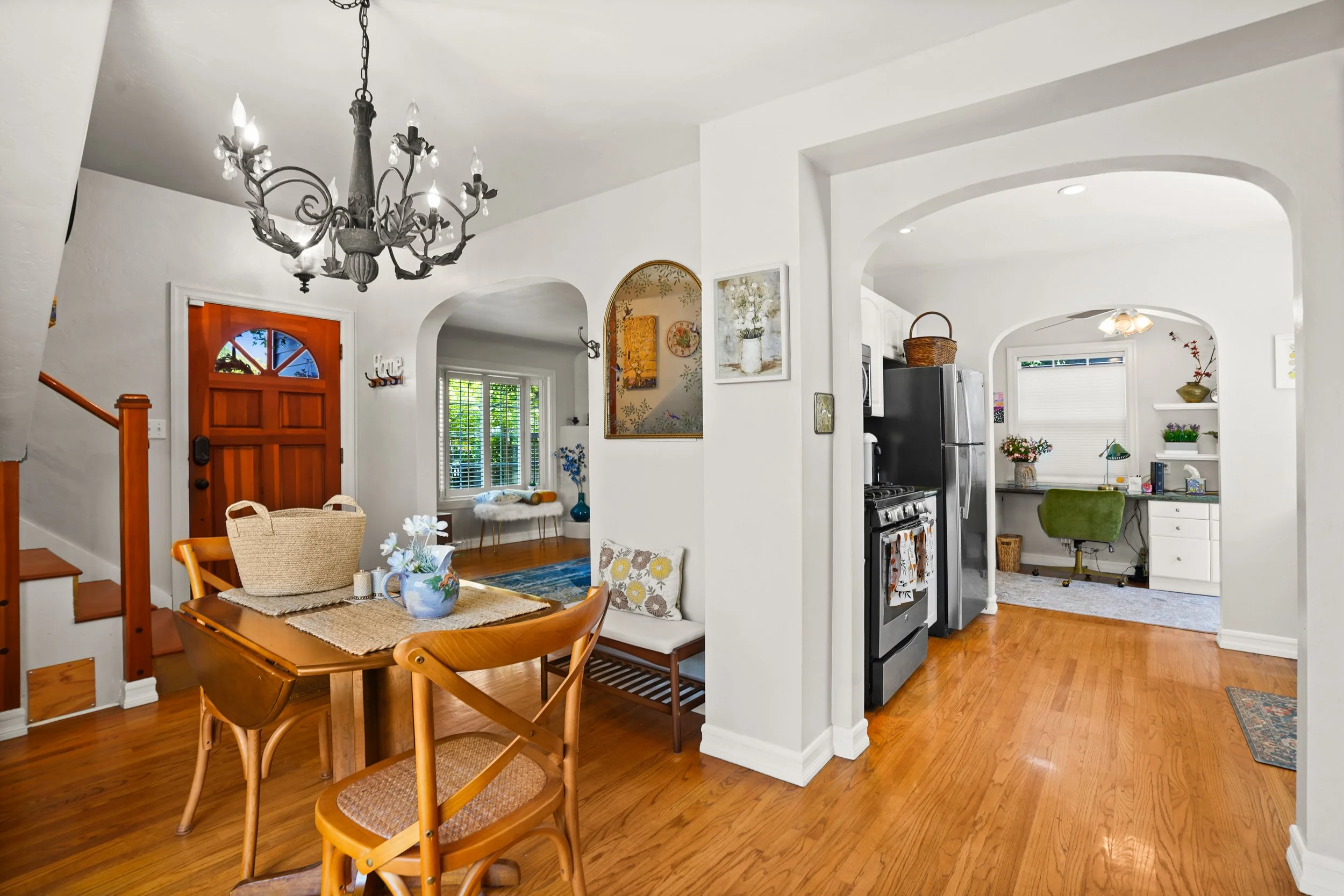 Inside view of a cozy home with a wooden dining table, chairs, and decorative paintings, leading to a kitchen with stainless steel appliances and white cabinets, brightened by natural light through windows.