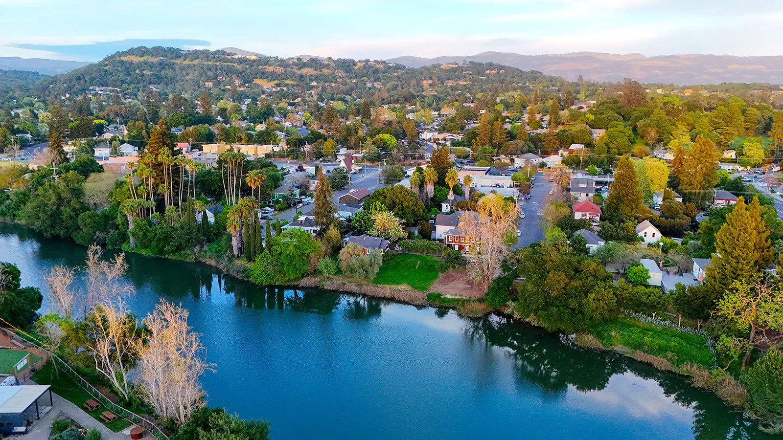 Aerial view of a small town by a river, with houses, trees, and hills in the background.