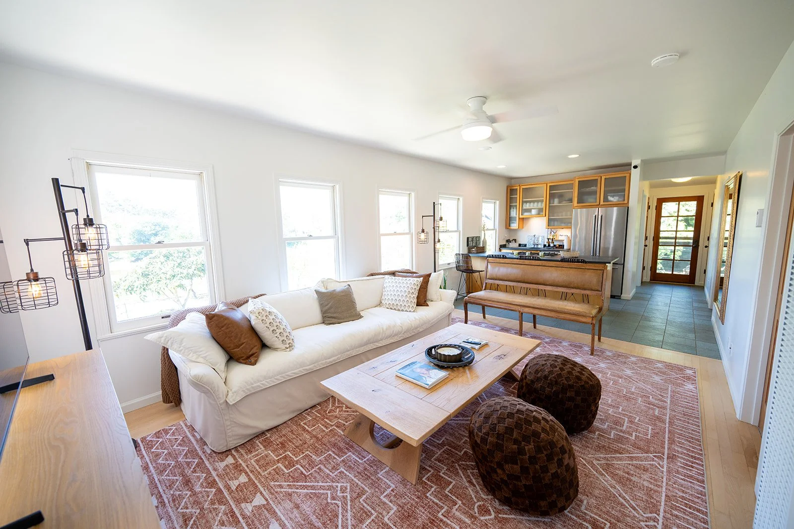 Bright living room with white walls, large windows, beige sofa with pillows, wooden coffee table, two dark patterned ottomans, modern floor lamp, and an open kitchen with wooden cabinets and stainless steel appliances at the back.
