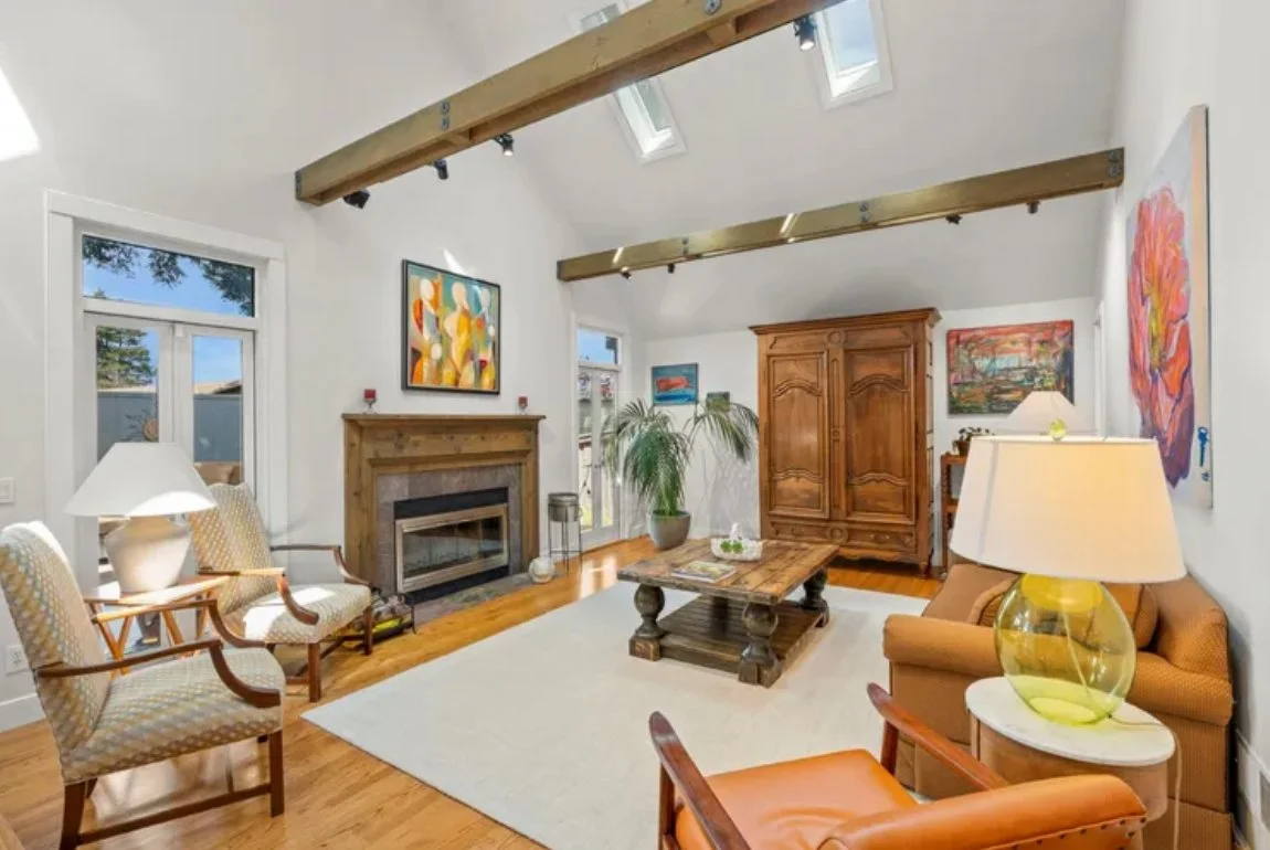 Living room with white walls, hardwood floors, and wooden ceiling beams. There are two skylights and three windows. The room features a fireplace with a wooden mantel, several colorful paintings on the walls, and a variety of seating including a beig