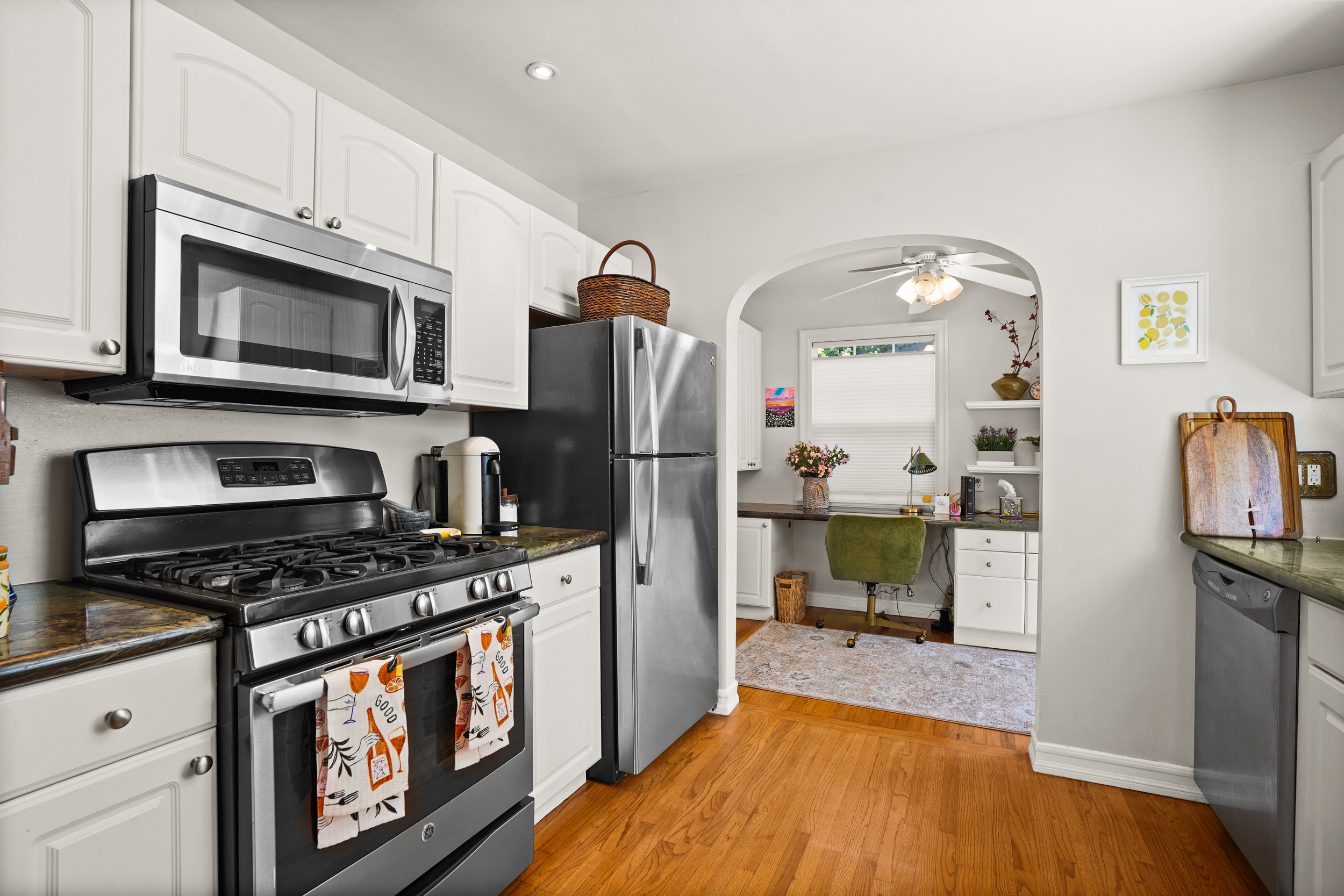 A kitchen with white cabinets, stainless steel appliances, a stove, microwave, fridge, and a small desk area with a green chair, decorated with plants and artwork.