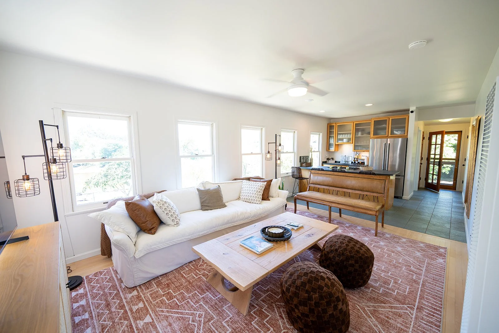 Open-concept living room and kitchen with white walls, a large white sofa, a light wood coffee table, patterned area rug, and multiple windows, leading into a kitchen with wooden cabinets and stainless steel appliances.