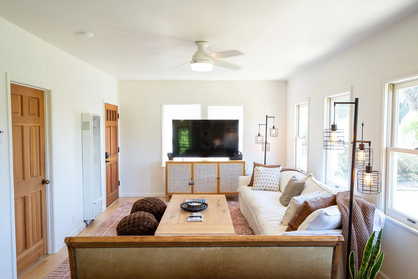 Living room with white walls, wooden doors, a large flat-screen TV on a cabinet, a beige sofa with pillows, a wooden coffee table, hanging light fixtures, and large windows letting in natural light.