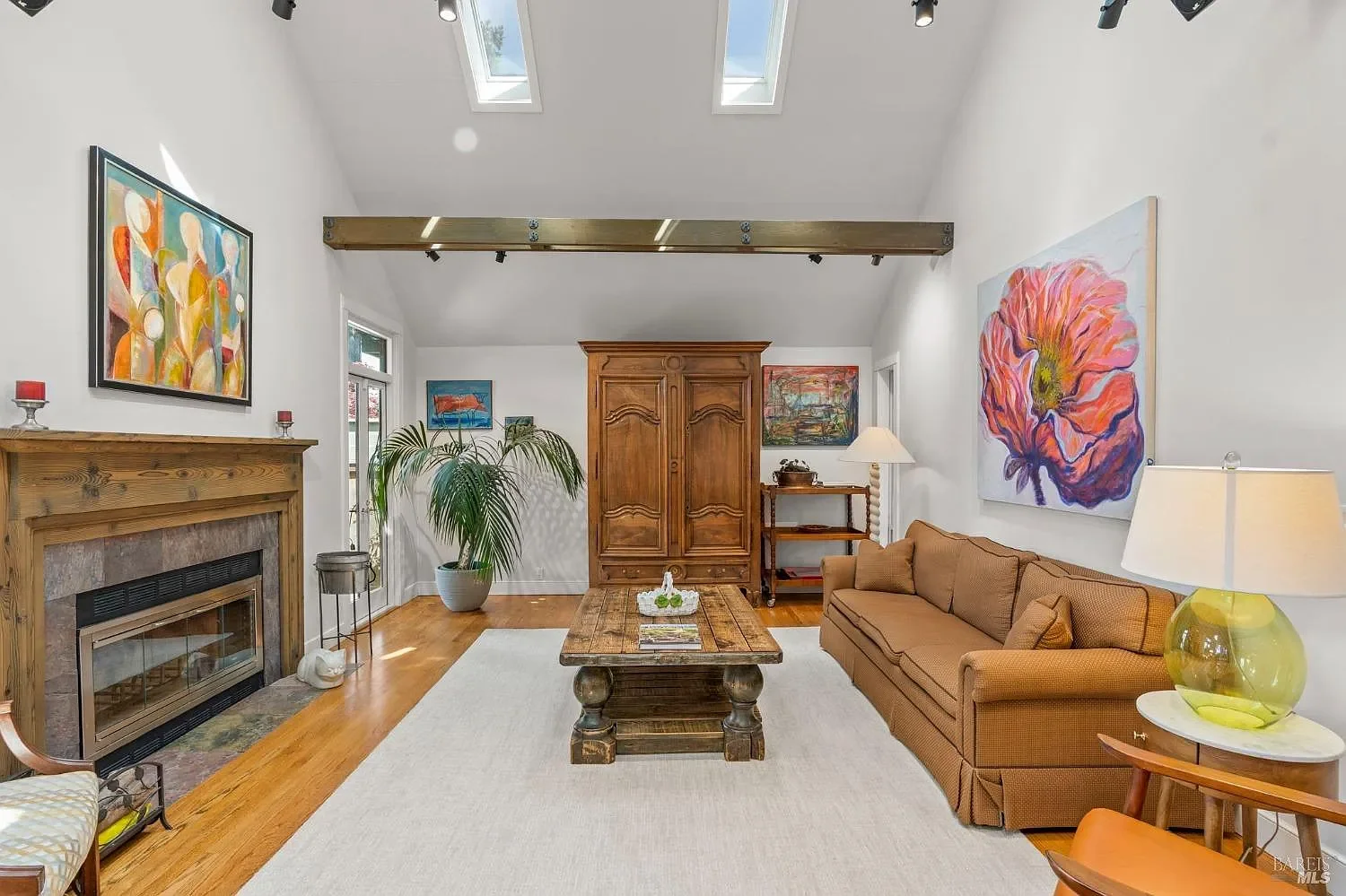 Bright living room with white walls and large ceiling skylights, featuring a neutral colored sofa, abstract floral artwork, and a wooden coffee table.
