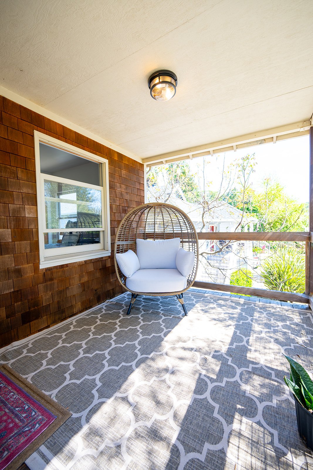 A cozy covered balcony with a white cushioned hanging chair, a patterned rug, a window on brown shingle siding, and a view of trees and neighbors outside.