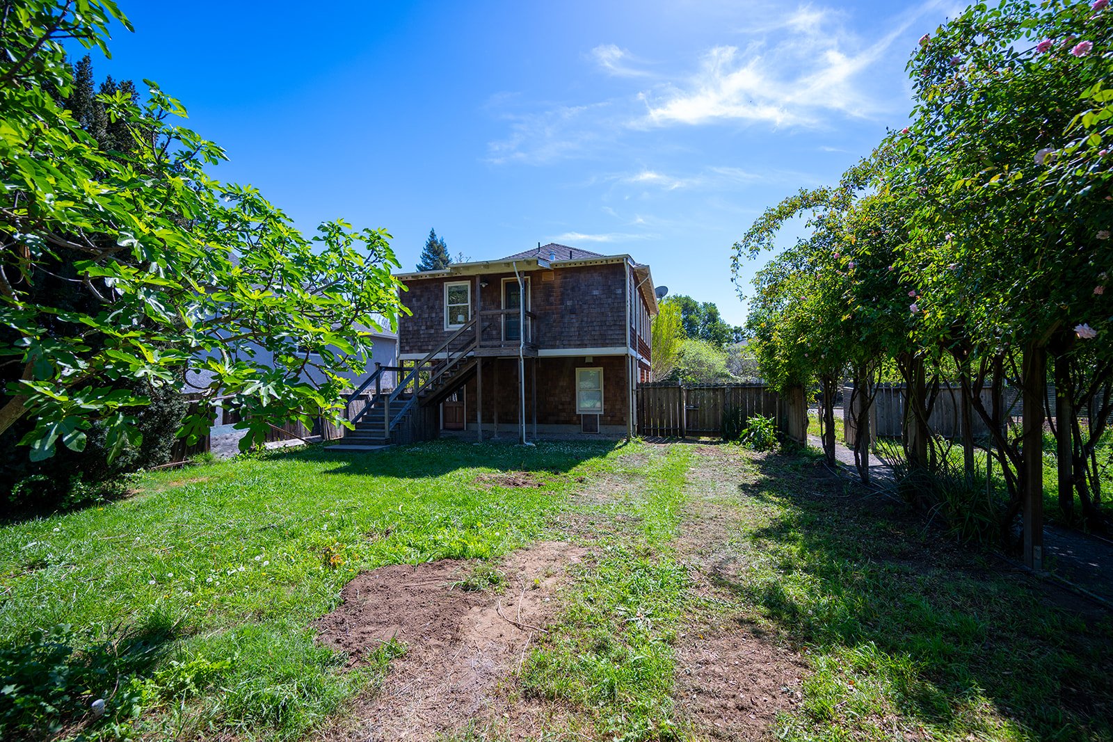 Backyard with grassy lawn, trees, and a wooden house with stairs and a second-floor balcony, under a clear blue sky.